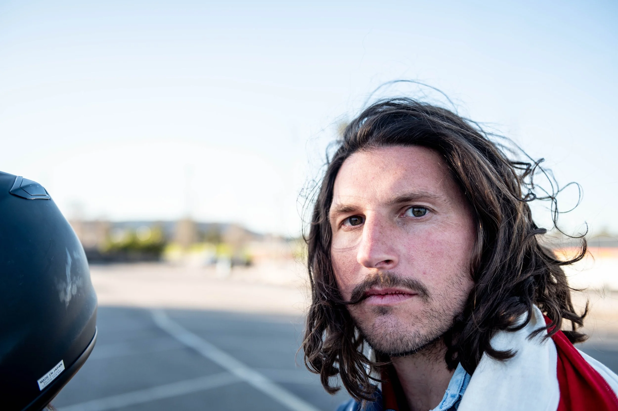 Close-up of a man with long, wavy brown hair and a beard, looking at the camera, with a parking lot and clear sky in the background.