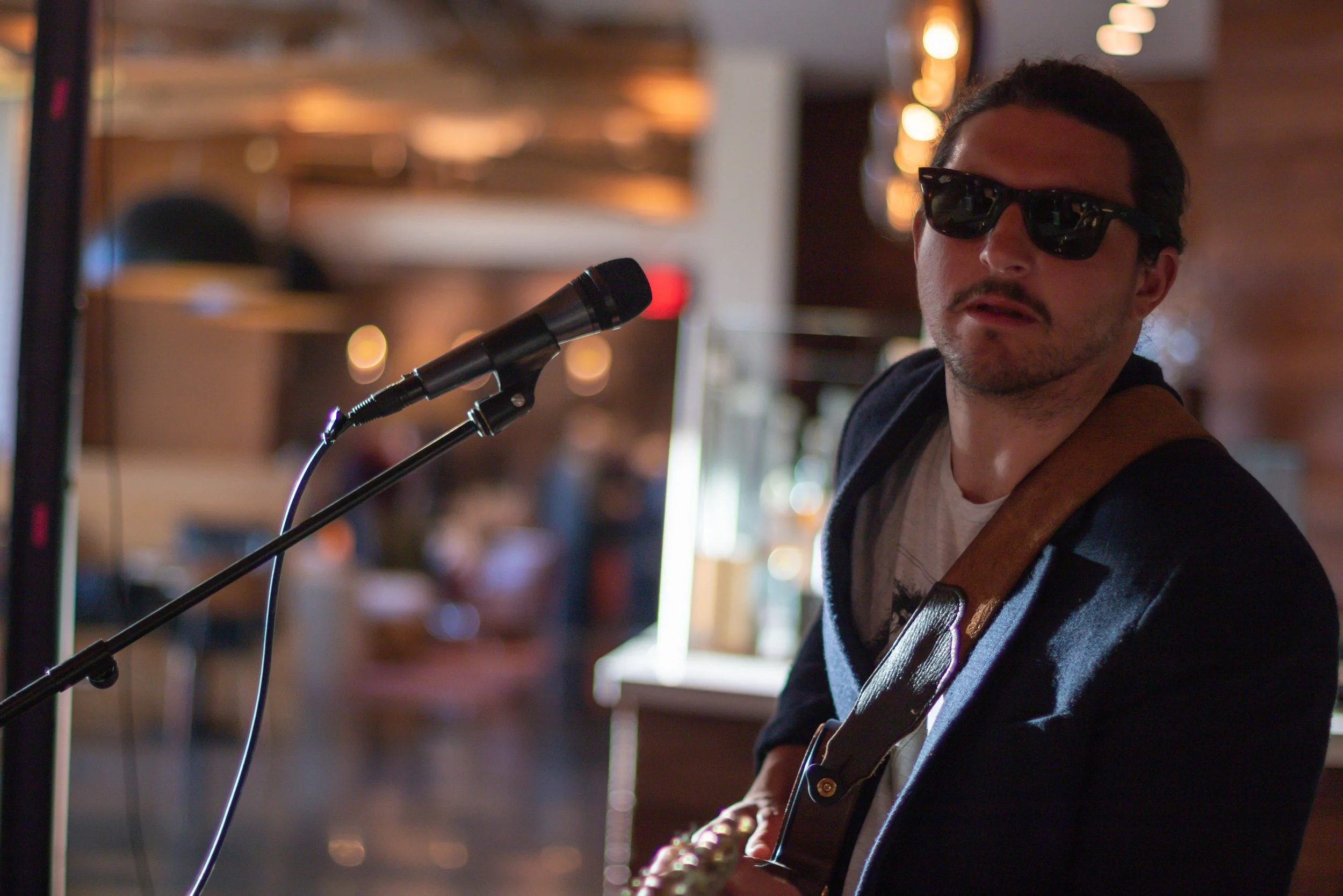 A man wearing sunglasses and a black jacket playing an acoustic guitar in a cozy indoor setting.