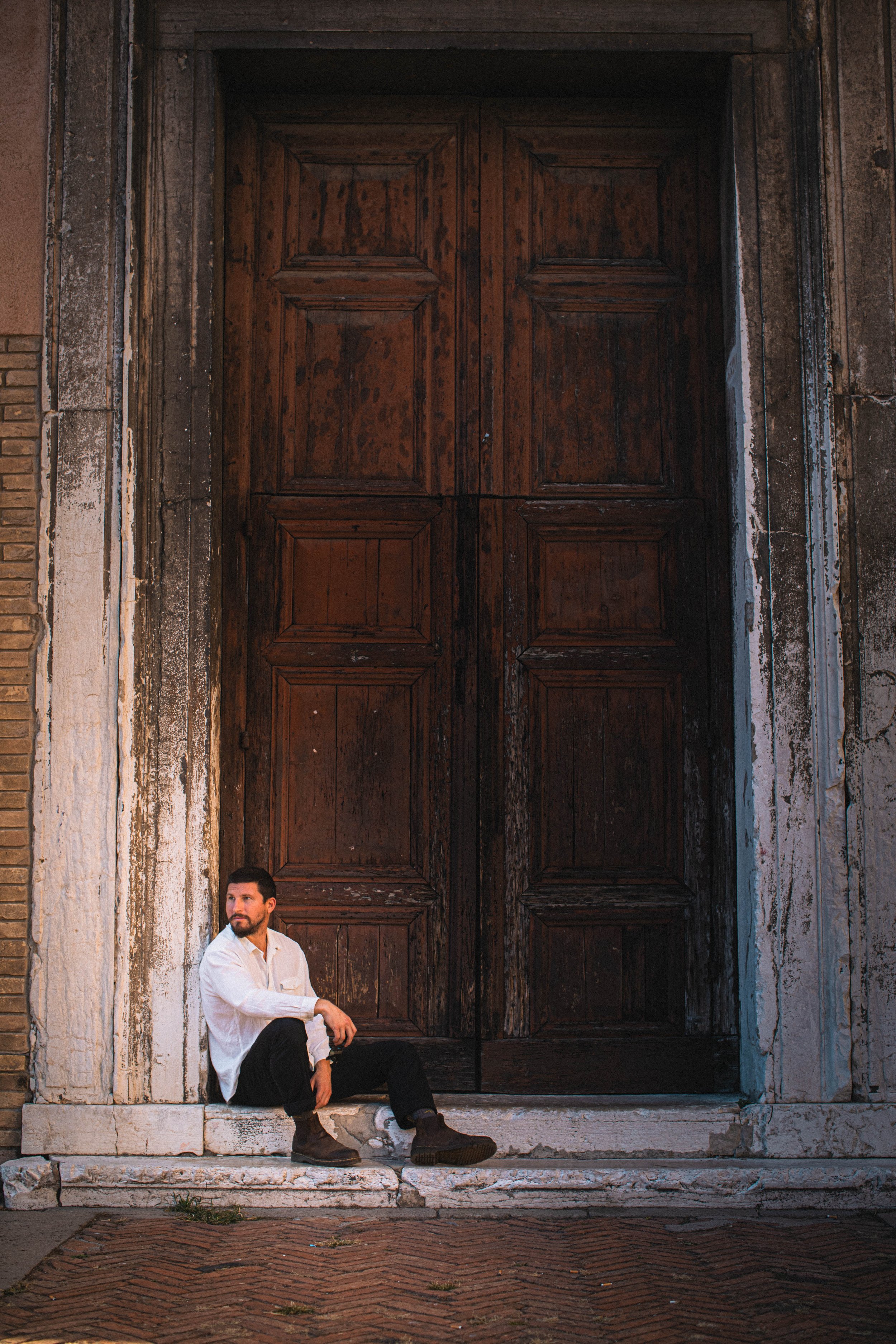 A man sitting on the steps in front of a large, old wooden door with decorative panels, set in a weathered stone building.