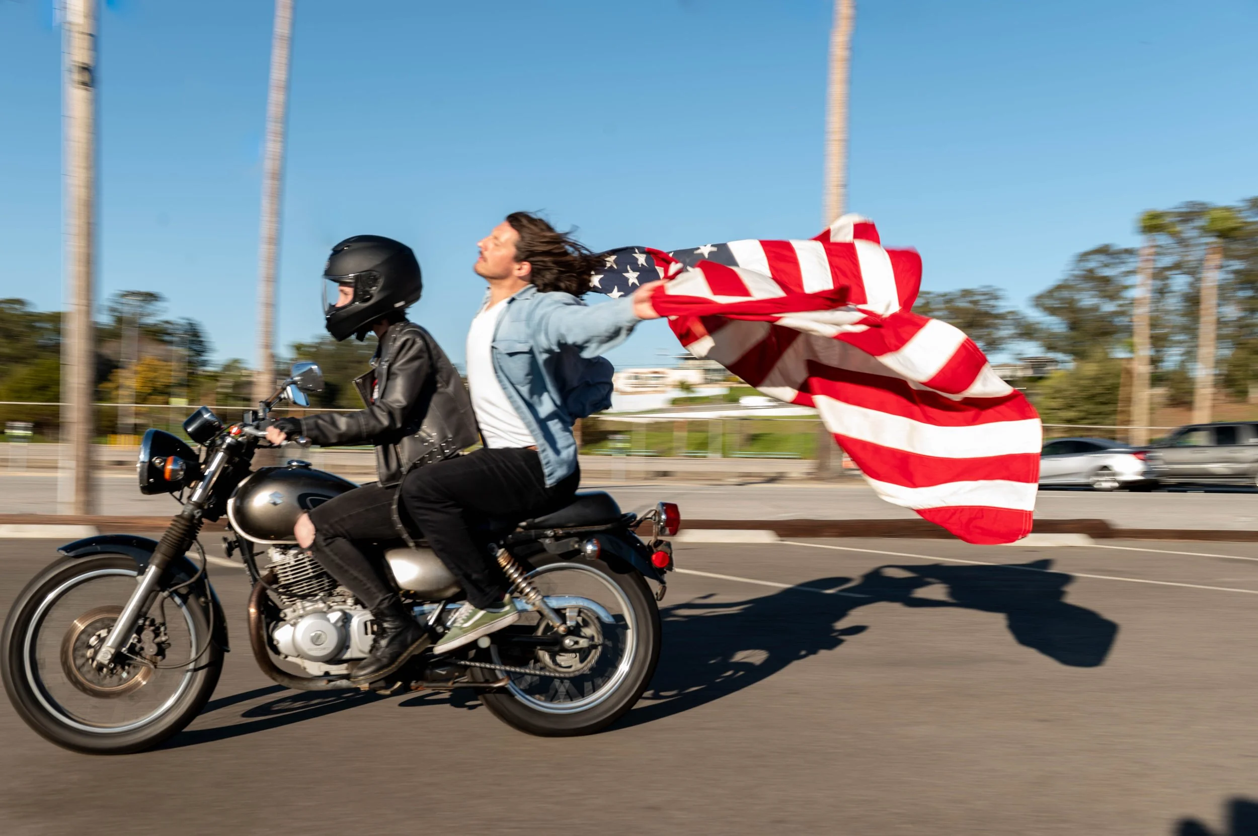 A person riding a motorcycle with a person holding an American flag attached to the bike on a road with trees and cars in the background.