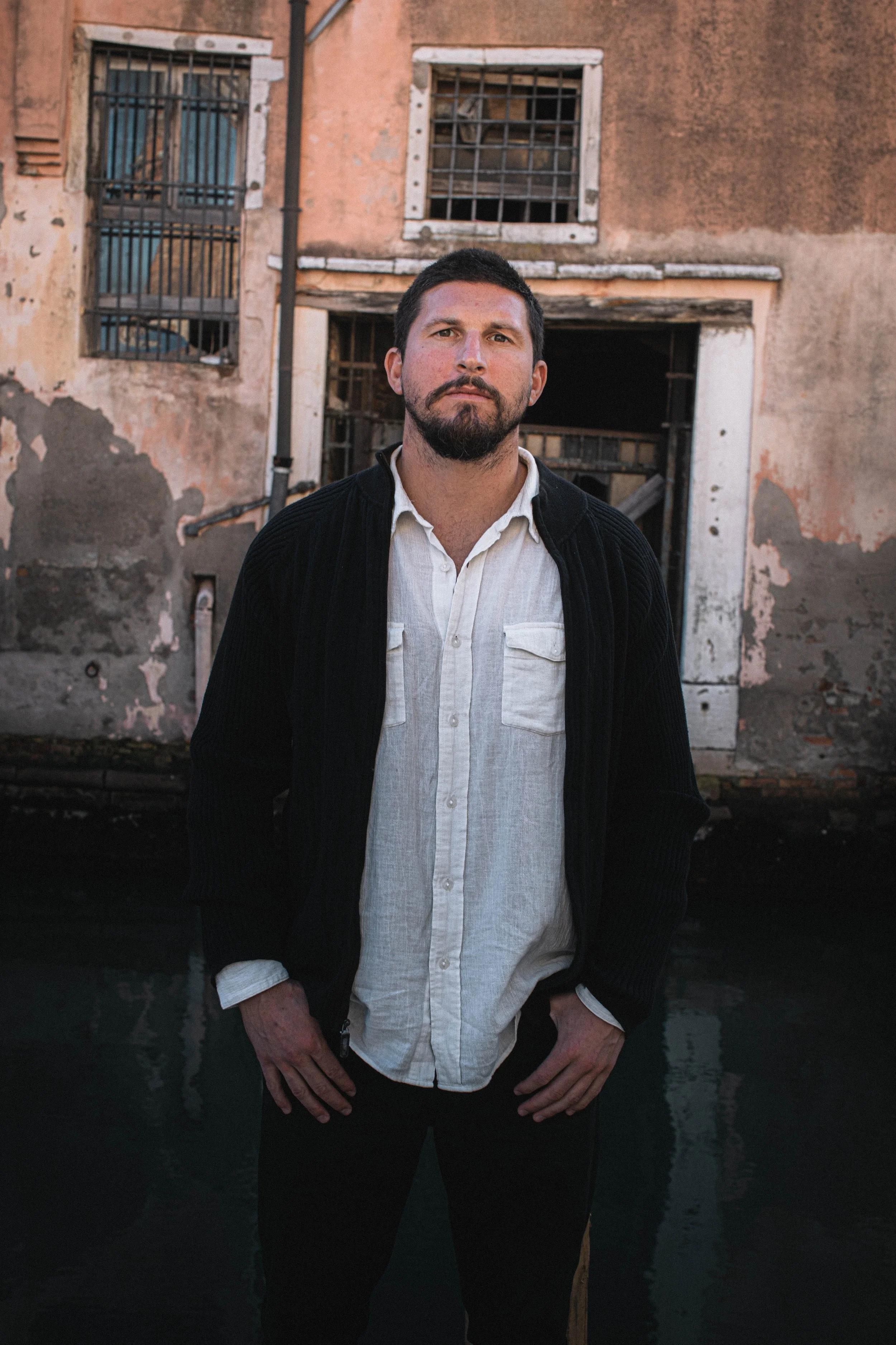 A man with dark hair and a beard standing in front of a weathered building with peeling paint and barred windows.