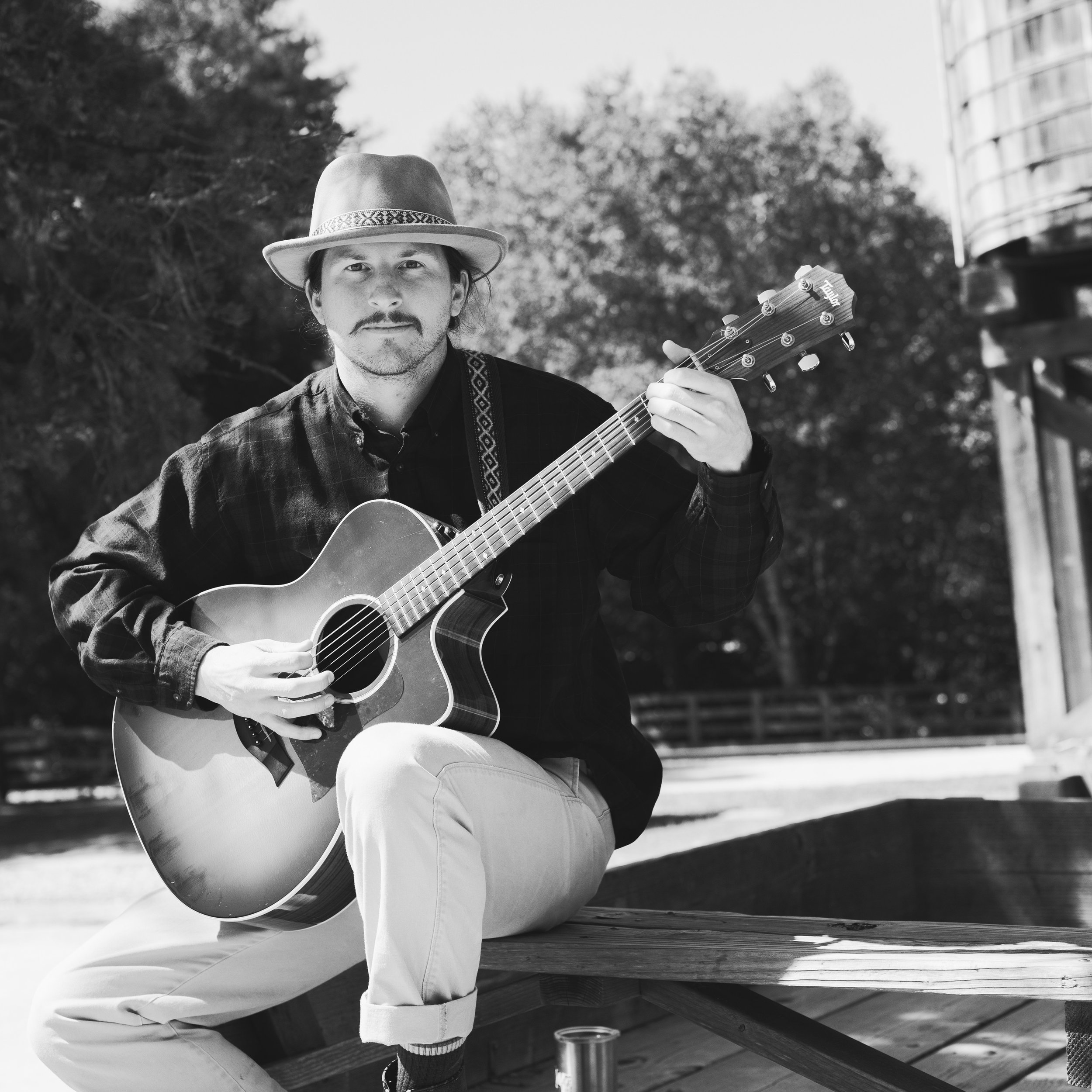 A man sitting outdoors on a wooden bench playing an acoustic guitar. He is wearing a hat, a plaid shirt, and light-colored pants, with trees and a building in the background.