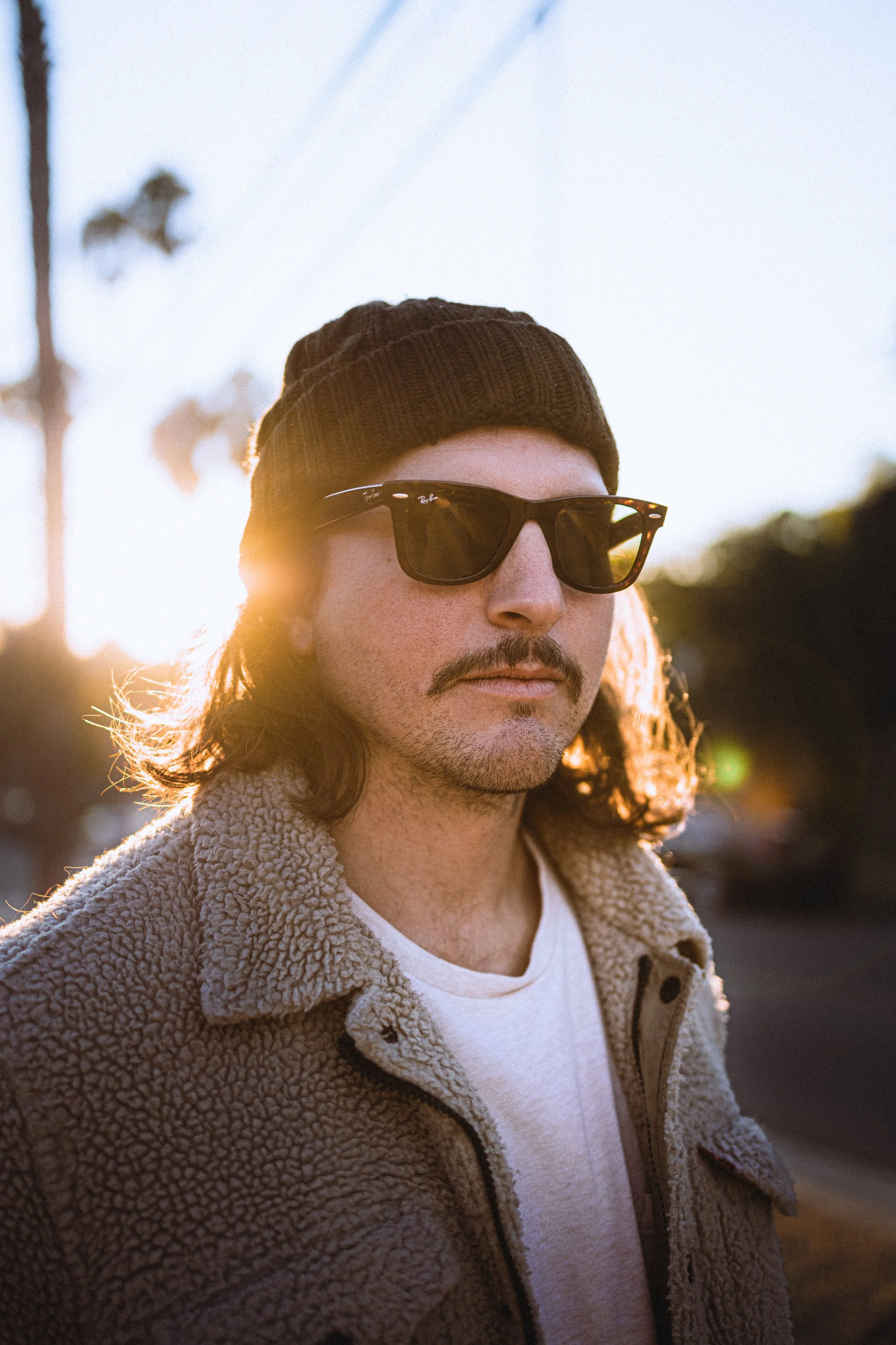 A man with long hair wearing a black knit beanie, sunglasses, a beige fleece jacket, and a white T-shirt, standing outdoors during sunset.