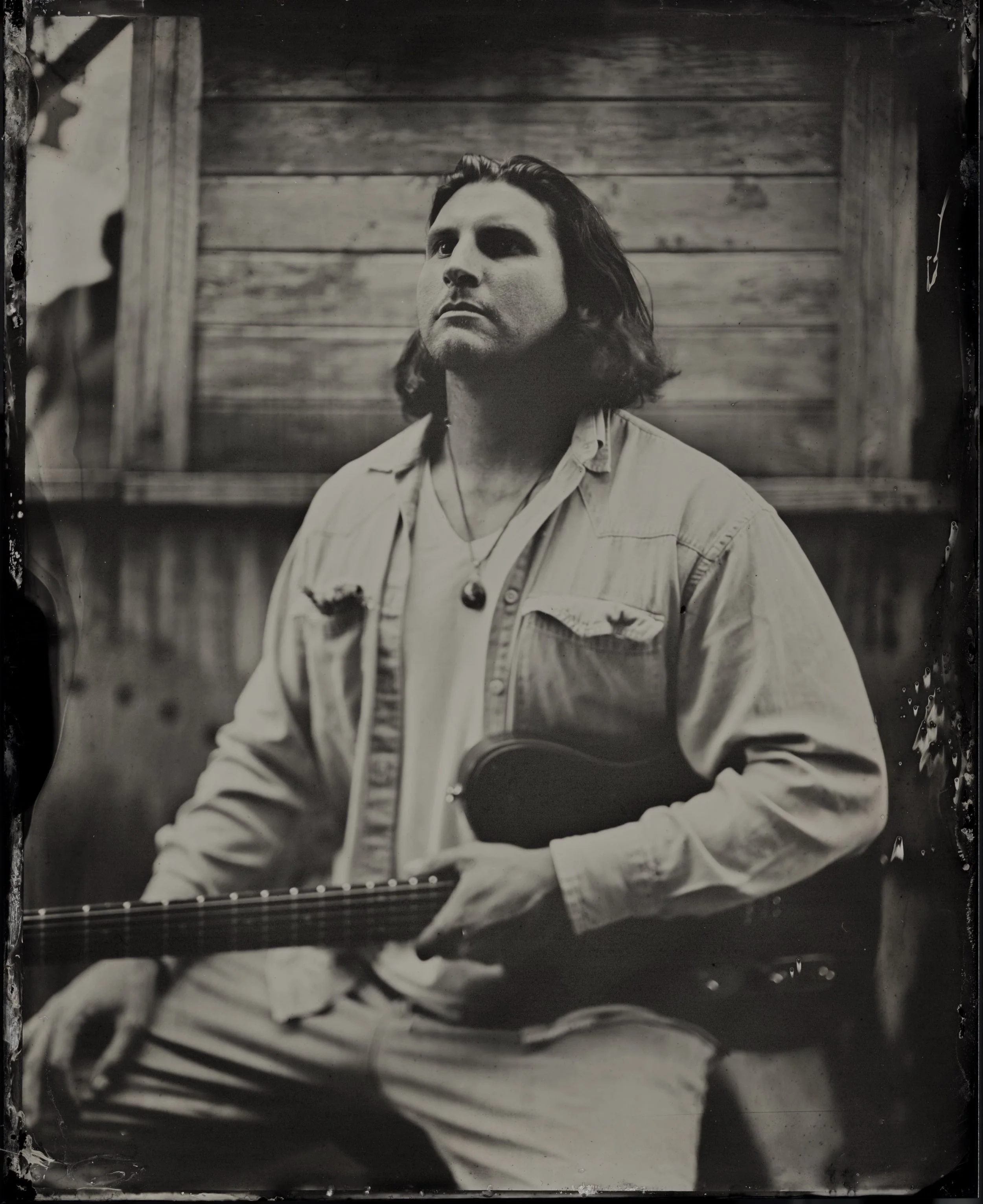 A black and white photo of a man with shoulder-length dark hair, wearing a light-colored shirt and a necklace, holding a guitar, standing in front of a wooden wall.