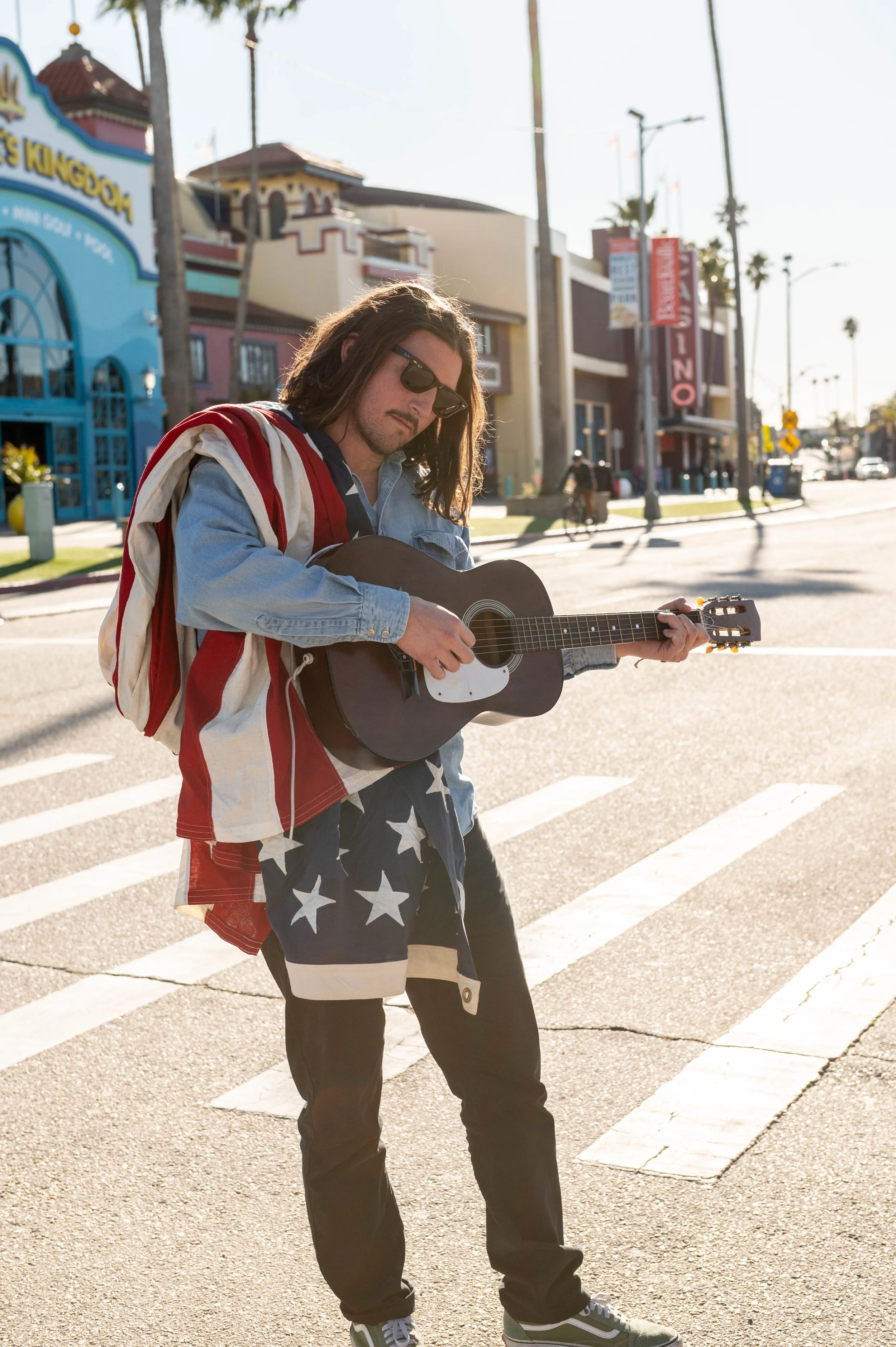 Young man with long hair playing guitar on a city street during sunset, wearing sunglasses, a denim jacket, and American flag-themed shorts.