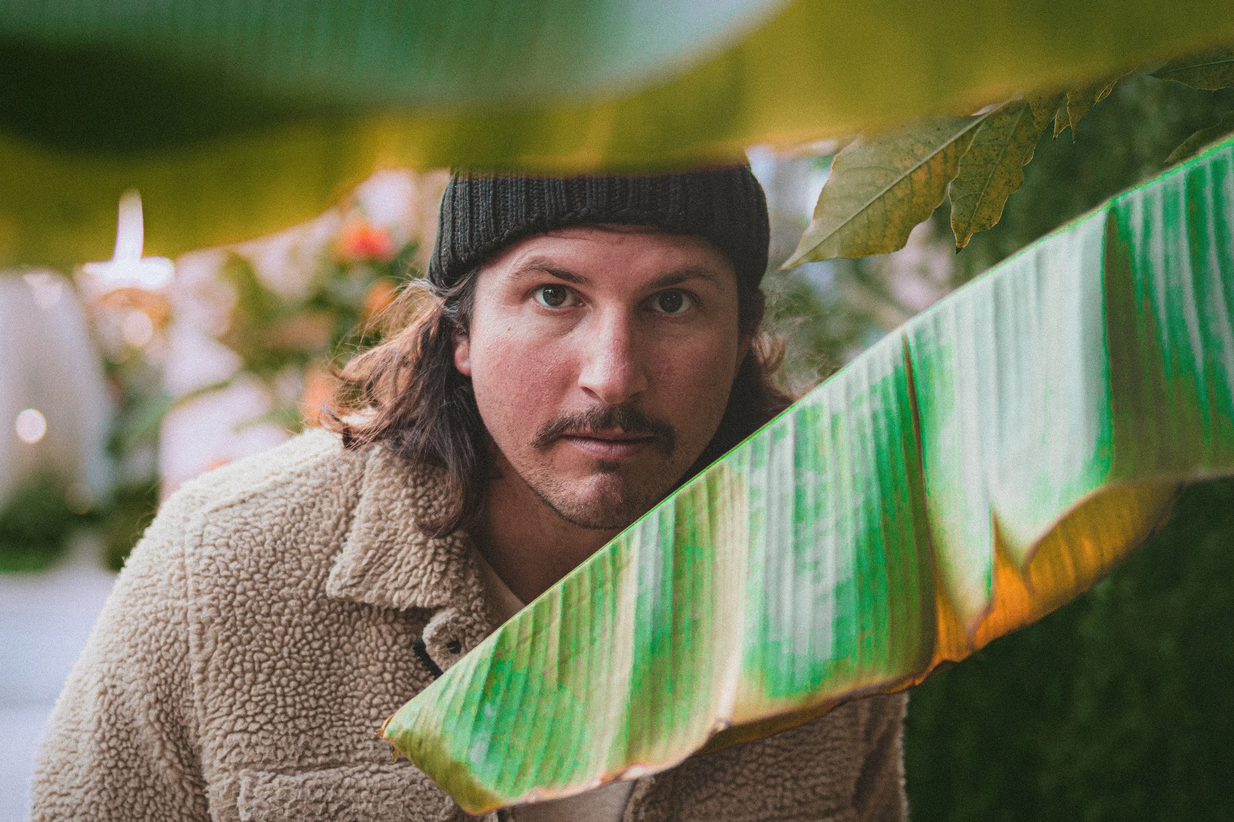 A man with long hair, beard, and mustache, wearing a dark knit beanie and beige fleece jacket, peering through large green leaves in a garden or jungle setting.