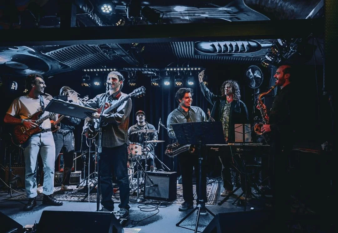 A band performing live on stage with musicians playing guitars, drums, keyboard, and saxophone, under stage lighting in a dark venue.