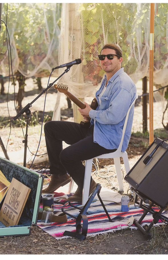 A man in sunglasses and a light denim shirt sitting on a white chair, holding an electric guitar, with a microphone stand nearby, outdoors with trees and a netted backdrop, surrounded by musical equipment and drinks on the ground.