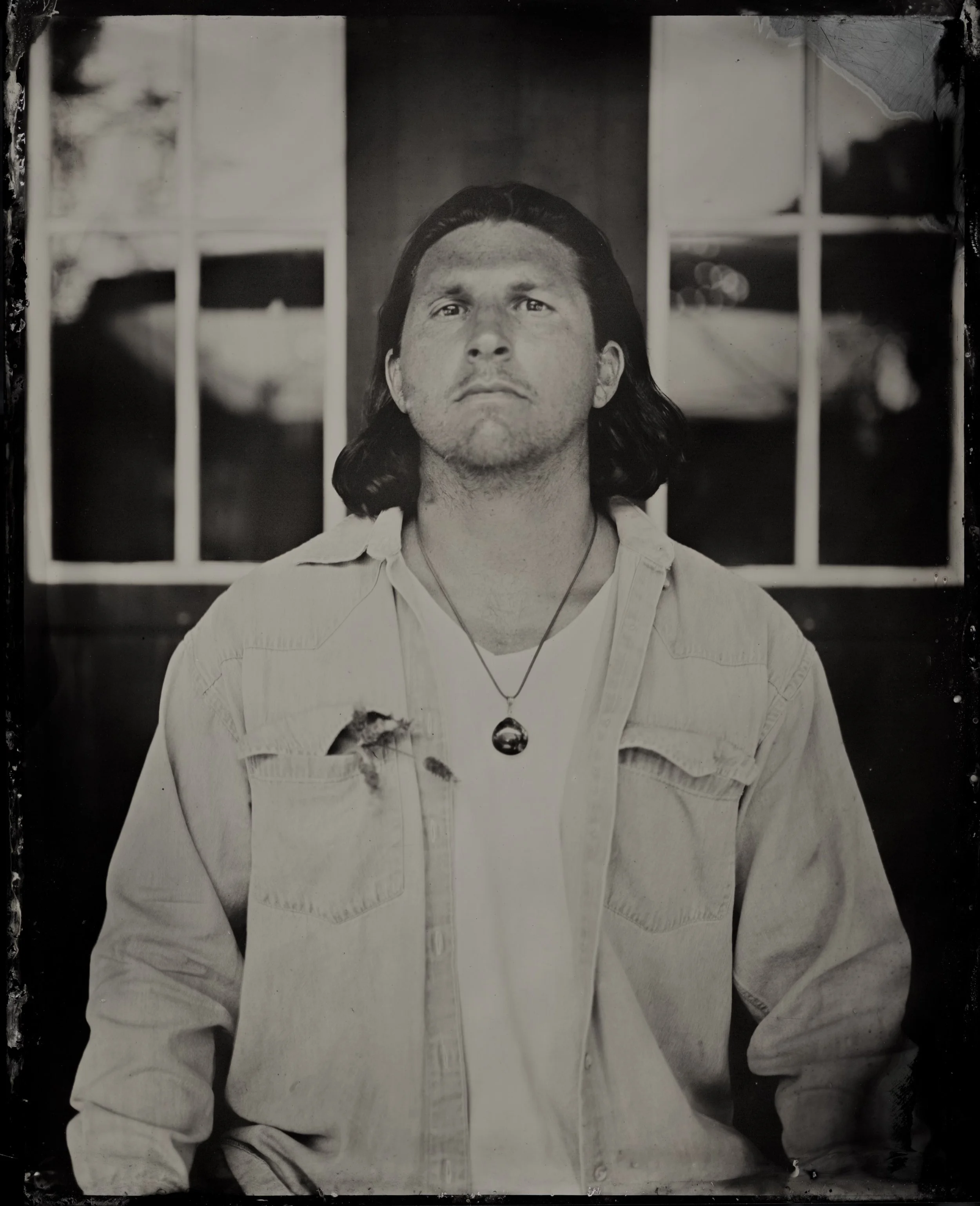 Black and white photo of a young man with long hair, wearing a light-colored jacket and a necklace with a pendant. He is staring directly at the camera with a serious expression, standing in front of a window with four panes.