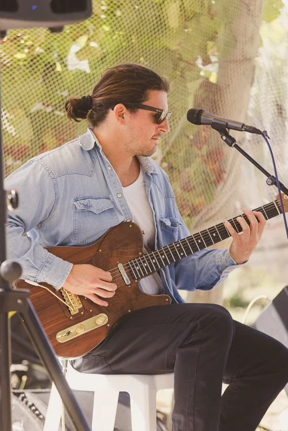 A man with long hair tied in a bun, wearing sunglasses and a light blue denim shirt, playing an acoustic guitar at an outdoor event with a microphone in front of him.