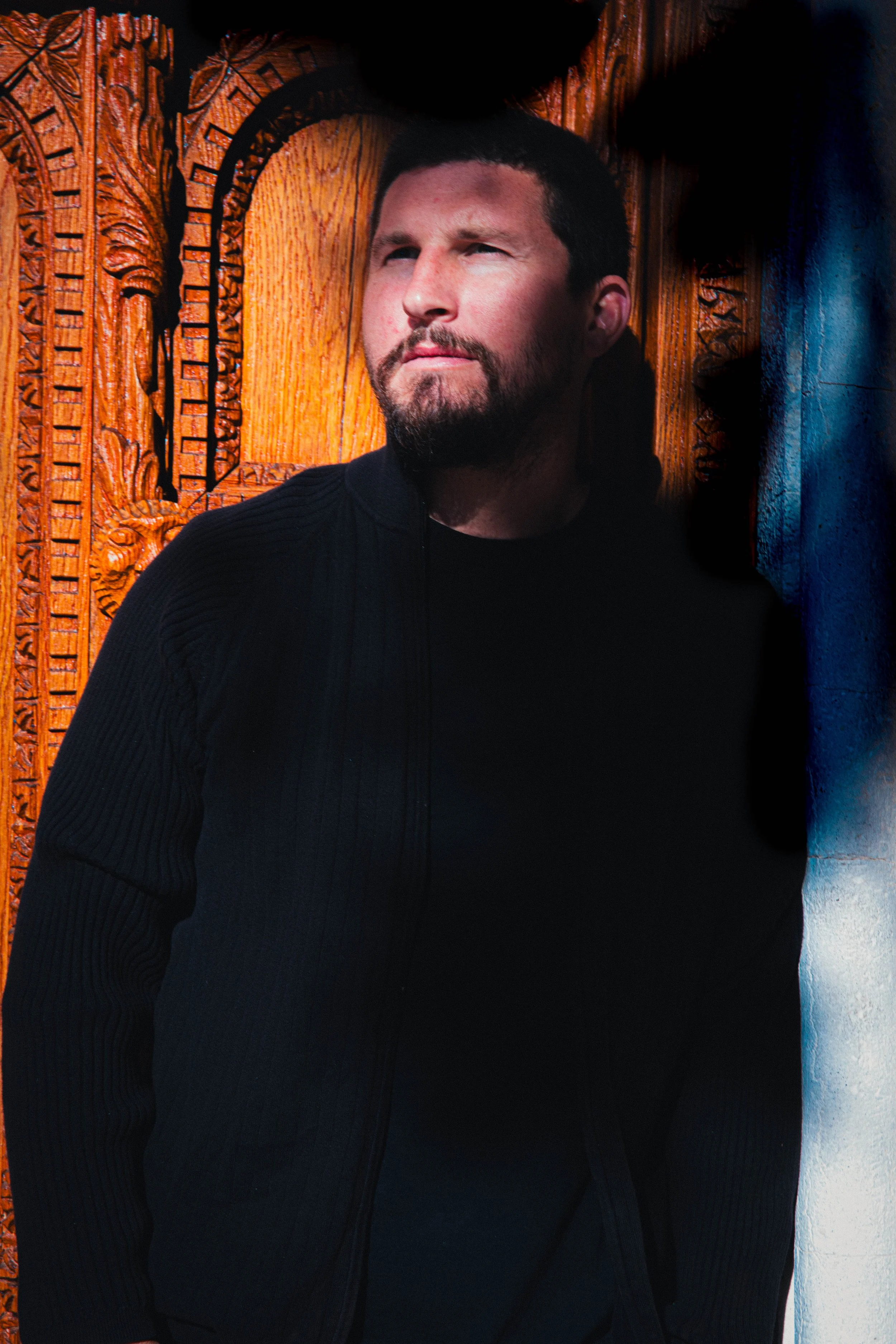 A man with a beard and short dark hair wearing a black zip-up sweater, standing in front of a carved wooden backdrop.