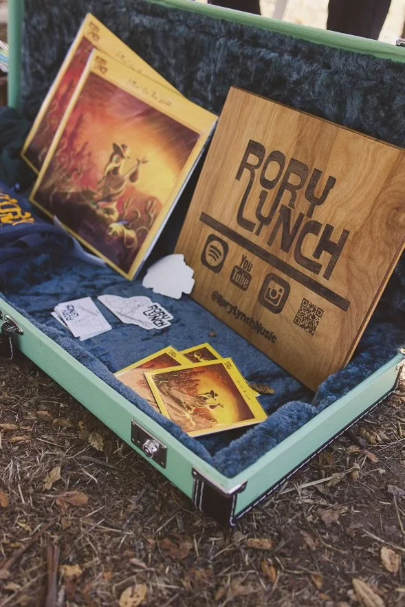 Open green suitcase with a dark blue interior, containing music promotional items and a wooden sign with the name Rory Lynch and social media icons.