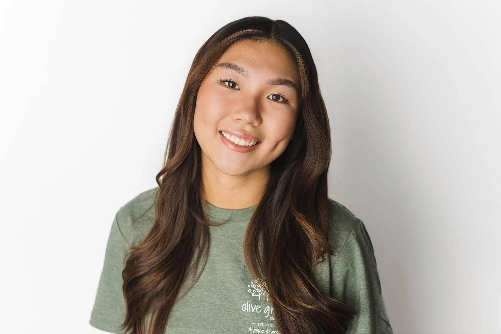A young woman with long brown hair, smiling, wearing a green T-shirt with white text, standing against a plain white wall.