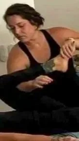 A woman with short dark hair wearing a black tank top appears to be giving a table Thai stretch in a room with light-colored walls and a striped blanket on the bed.