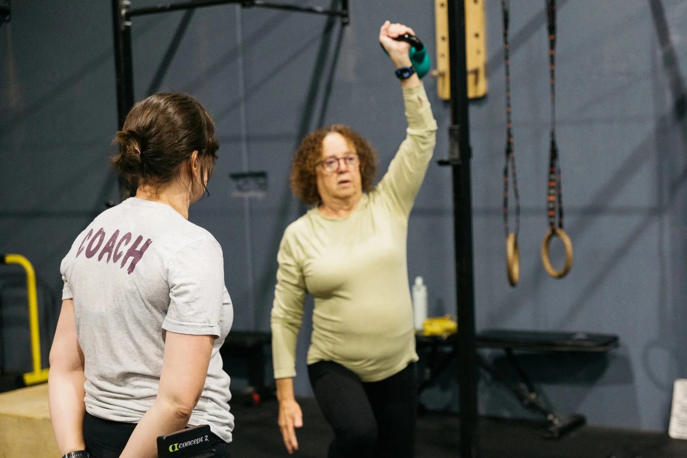 Women lifting a barbell in a gym, with a trainer assisting.