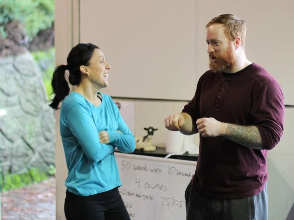 A woman and man are talking and smiling indoors, with a whiteboard visible in the background.