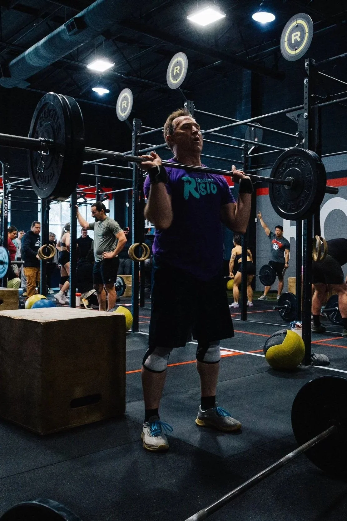 A man lifting a barbell during a CrossFit workout in a gym filled with other athletes and workout equipment.
