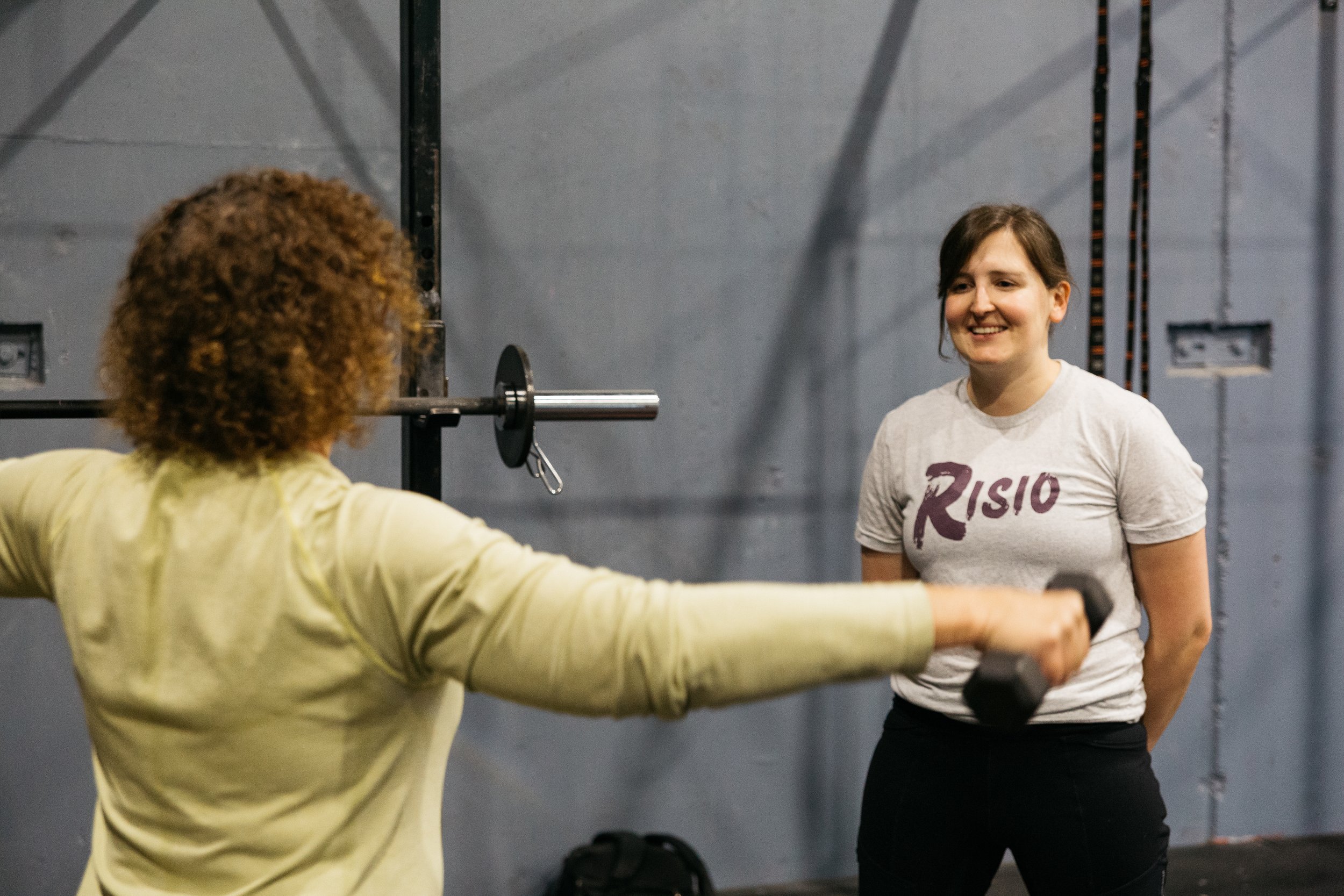 Two women in a gym, one with curly hair and a yellow shirt, and the other with brown hair, a gray T-shirt, and black gloves, are standing close to each other, smiling, and talking. The woman with curly hair has her arms extended to the sides.