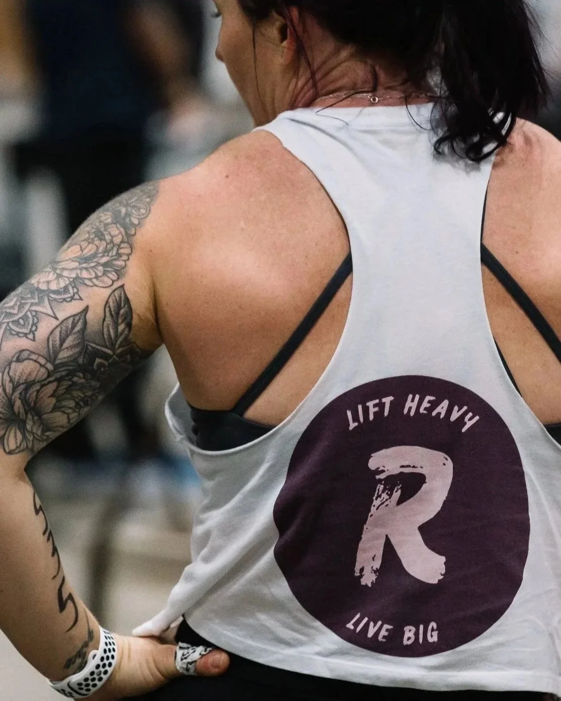 Close-up of a woman in a white athletic tank top with a logo that says 'Lift Heavy Live Big' and a large 'R.' She has tattoos on her left arm and is wearing black sports bra straps underneath.