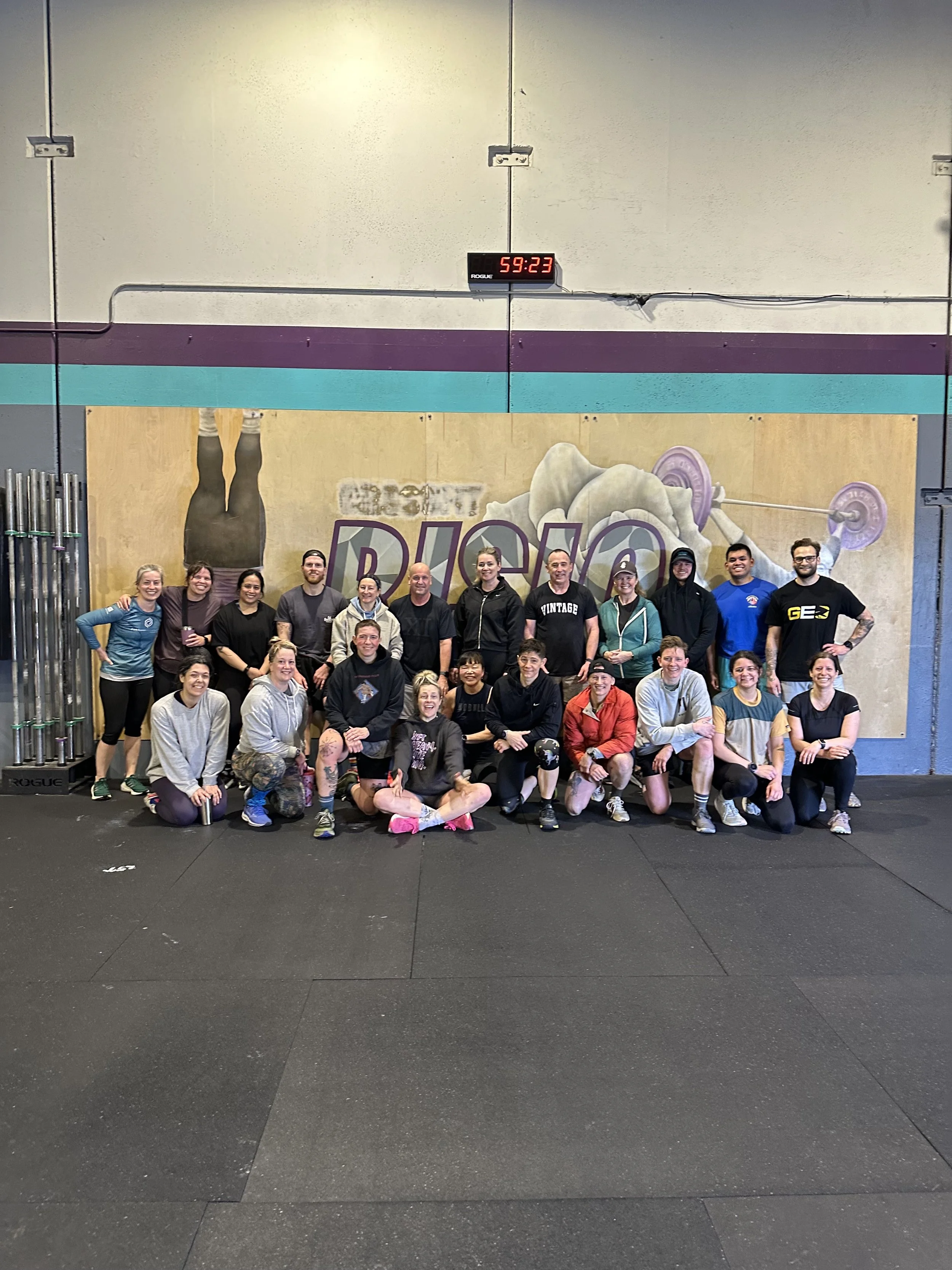 A group of people posing for a photo in a gym or CrossFit gym, with a large mural of a person lifting a barbell on the wall behind them.