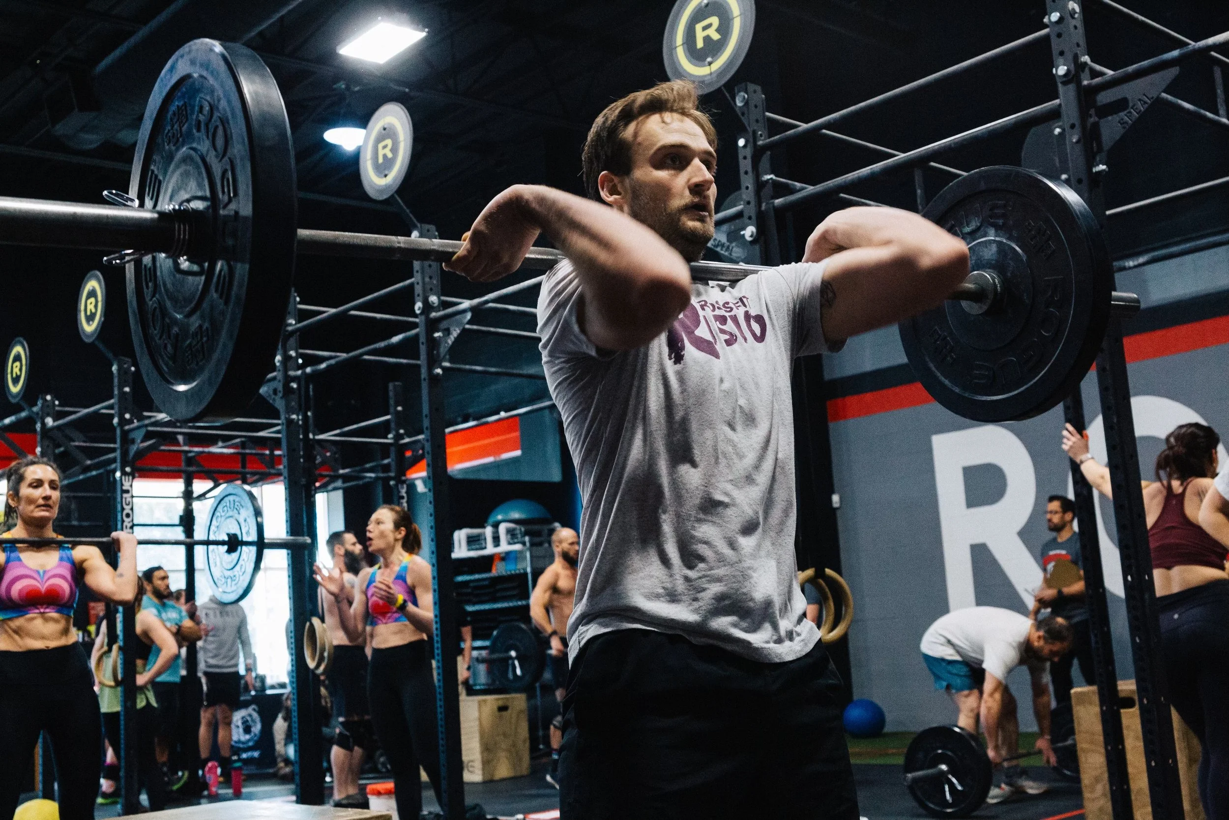 A man lifting a barbell during a CrossFit workout at a gym filled with other people also working out, with CrossFit equipment and branding visible.