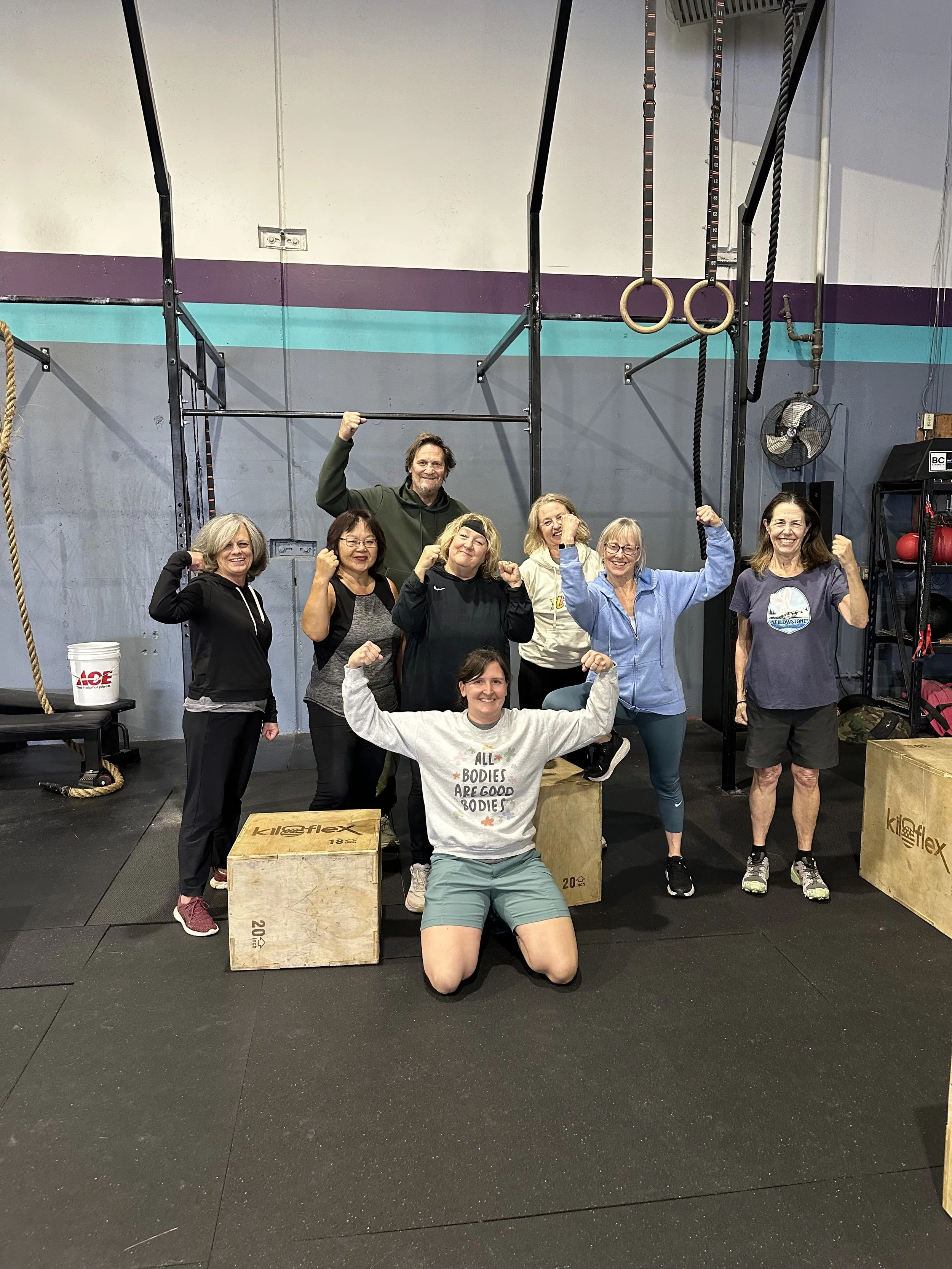 A group of nine women and one man at a gym posing with fighter fists, some flexing their arms, in front of wall-mounted gymnastic rings, ropes, and a fan, with wood plyometric boxes in the foreground.