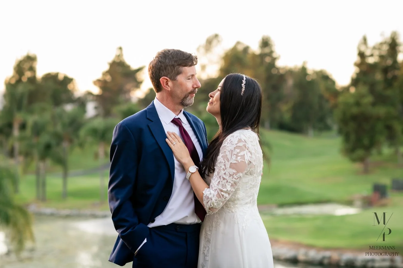 A bride and groom standing closely outdoors, gazing into each other's eyes, with a scenic park and water in the background during sunset.