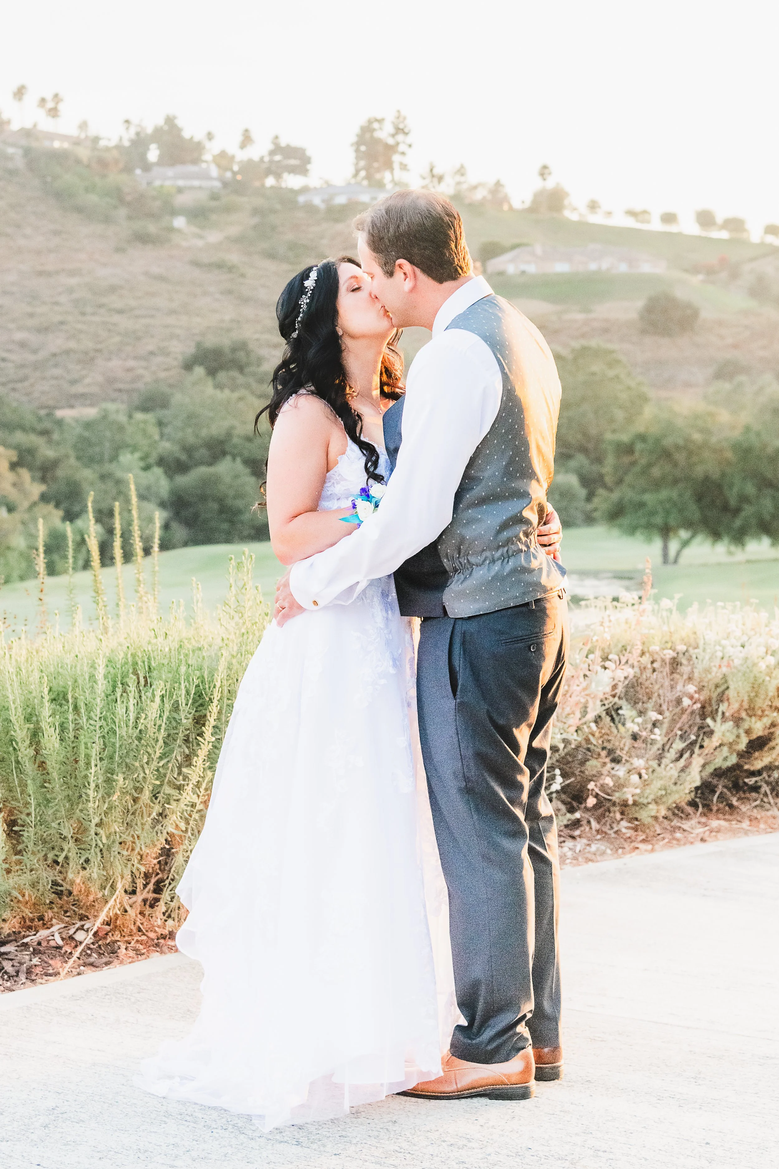A bride and groom are kissing outdoors during sunset on a hillside with houses and trees in the background.