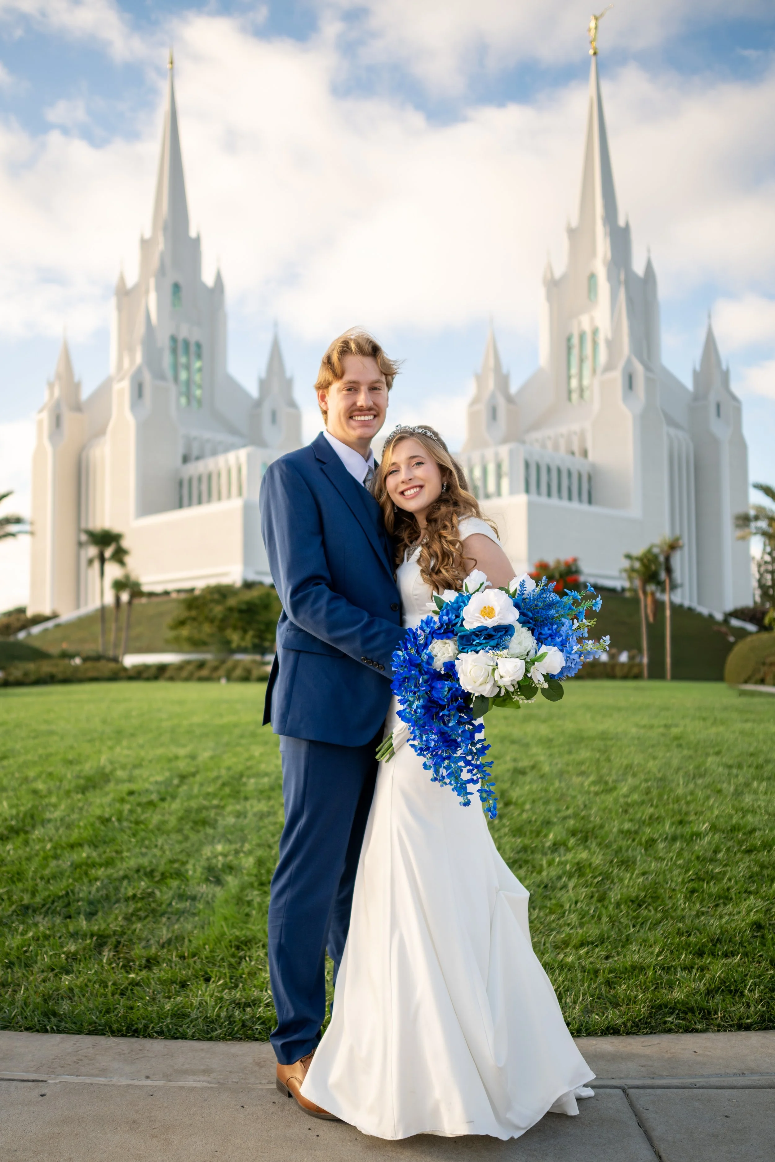 A smiling bride and groom standing on a sidewalk in front of a white church, with the groom in a blue suit and the bride in a white wedding dress holding a large bouquet of blue and white flowers.
