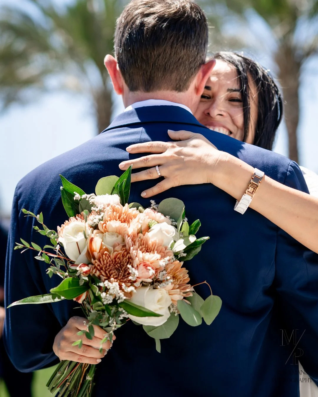 A man and woman embracing outdoors, with the woman smiling and holding a bouquet of flowers, the woman's hand with a wedding ring visible.