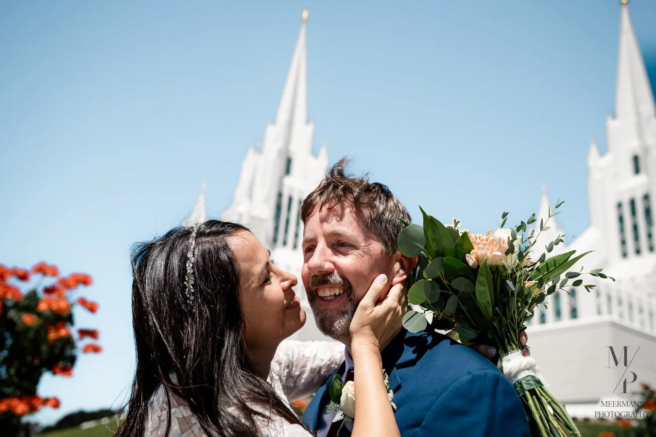 A bride and groom sharing a close moment outdoors, with a church in the background, during their wedding. The bride is holding a bouquet of flowers, and both are smiling.