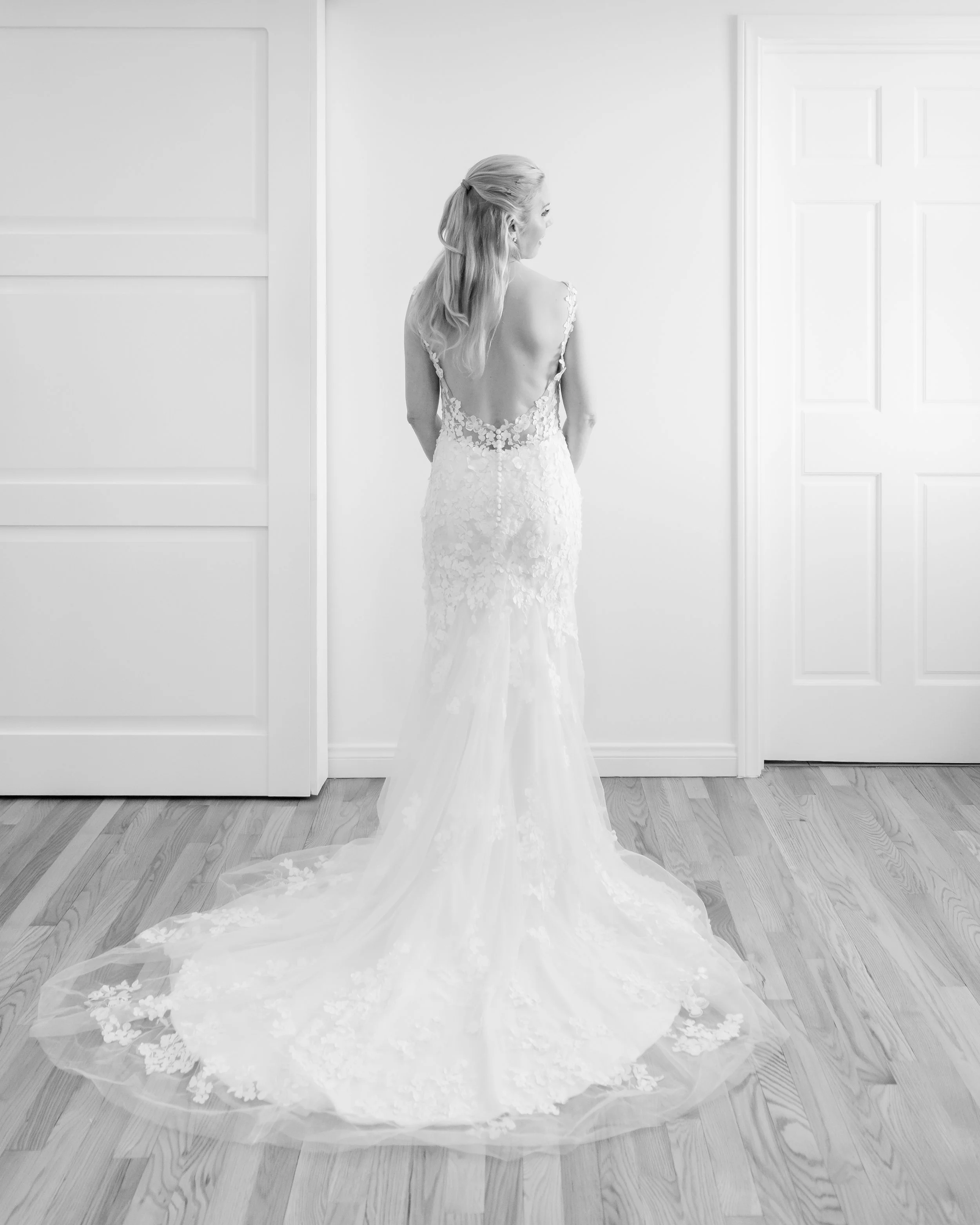 A bride in a wedding dress with a low back and floral lace details standing on a wooden floor, with a train extending behind her, in a room with white walls and doors.