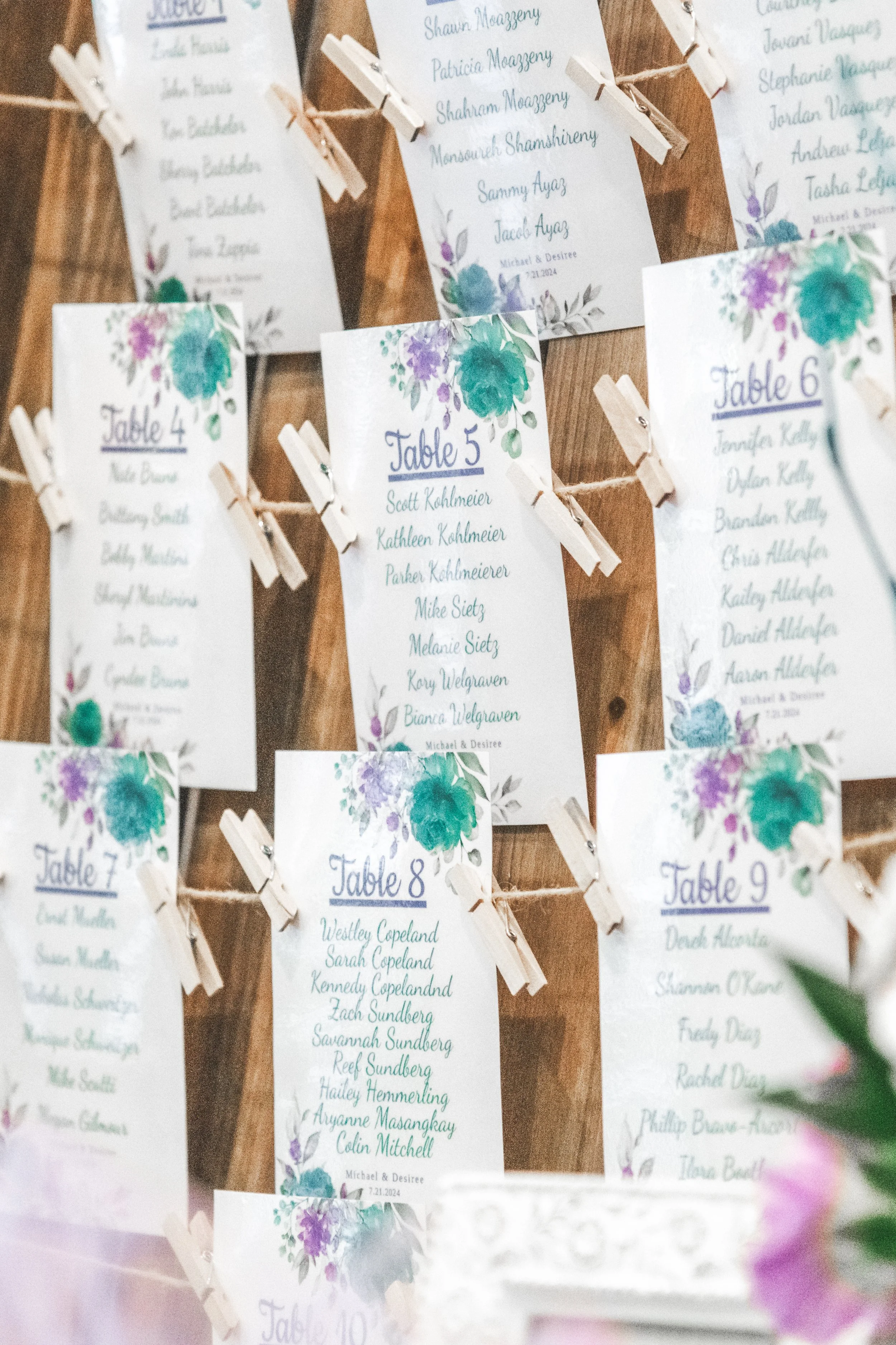 A display of wedding table assignments printed on white cards with purple and teal floral accents, hanging from strings with miniature clothespins. The cards are labeled with table numbers and list guest names.