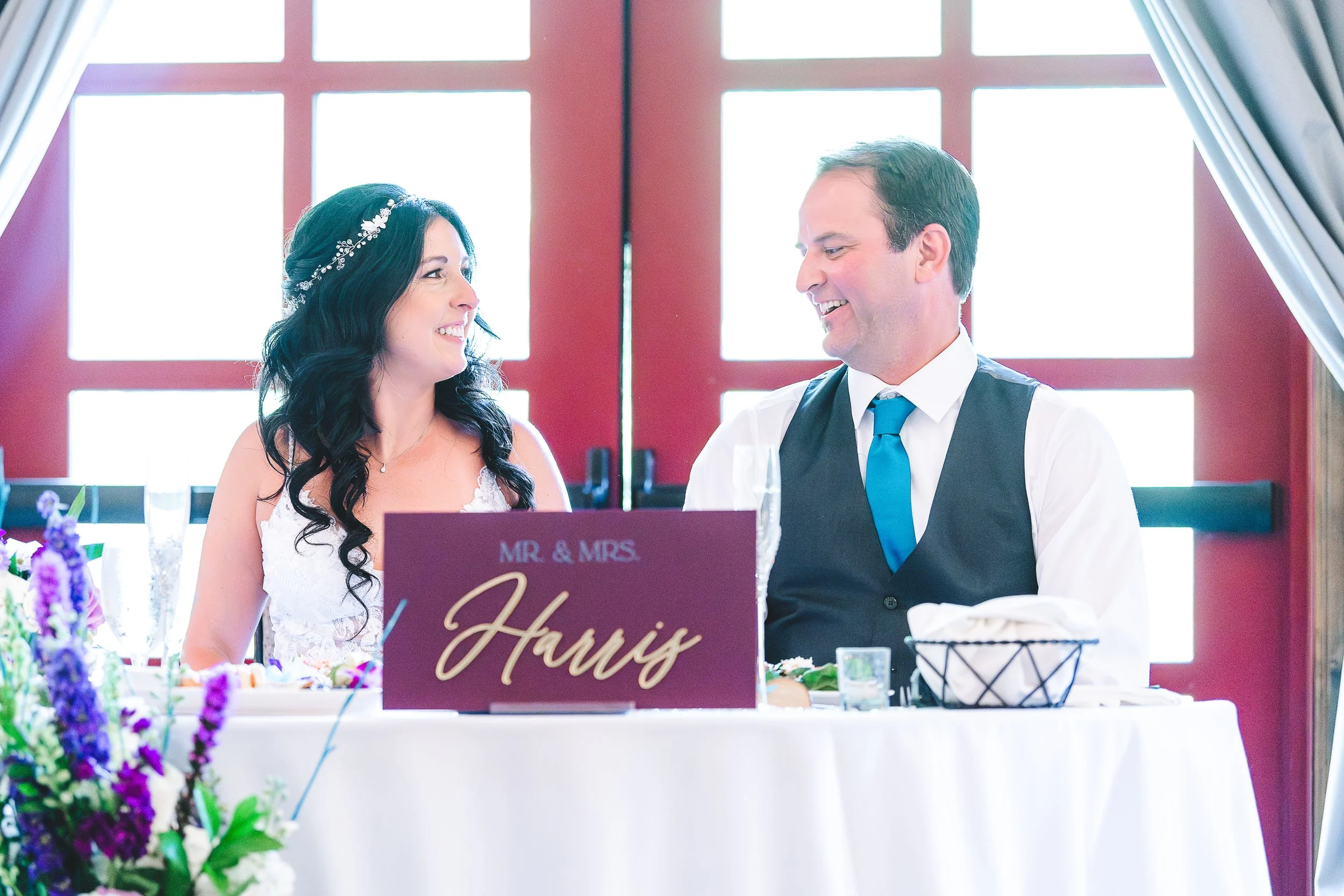 A smiling bride and groom sit across from each other at a wedding reception, with a sign that says 'Mr. & Mrs.' in front of them. The bride has long dark hair with a floral headpiece, and the groom is dressed in a white shirt, black vest, and blue ti
