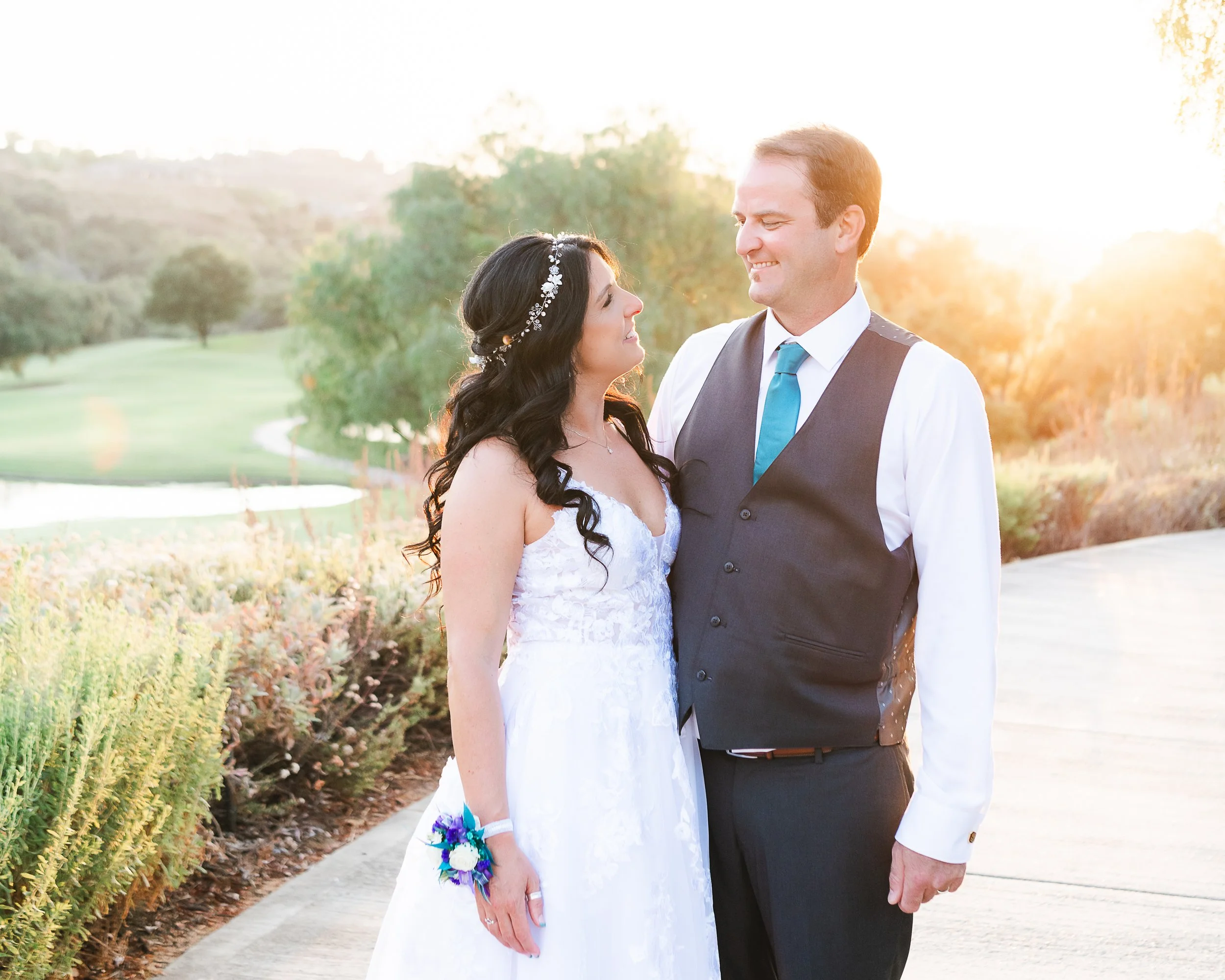 A bride and groom stand close together outdoors during sunset, smiling at each other. The bride wears a white wedding dress and a floral headband, holding a small bouquet. The groom wears a dark vest, white shirt, and teal tie. Background features tr