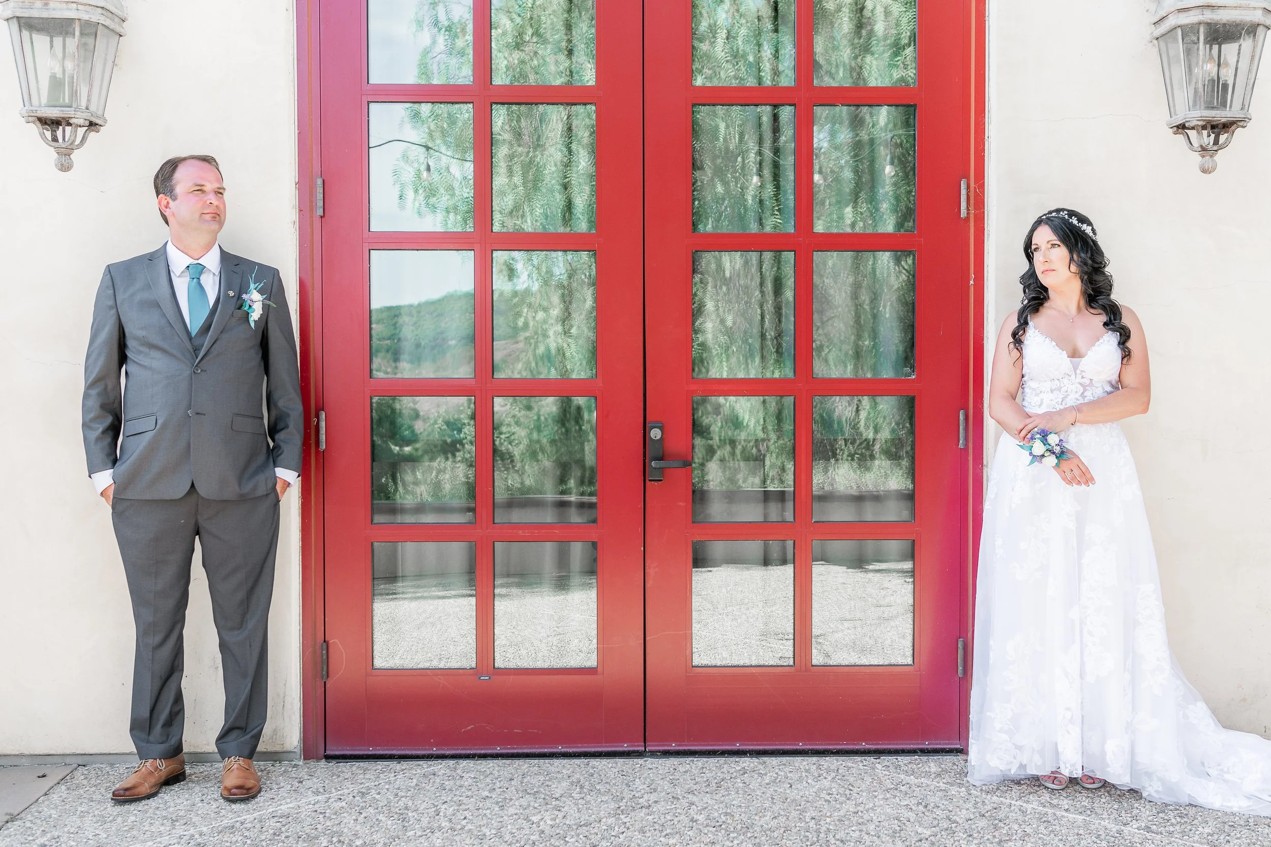A bride and groom standing on either side of a red door with large glass panes, outdoors. The groom is wearing a gray suit and teal tie, and the bride is in a white wedding gown holding a bouquet.