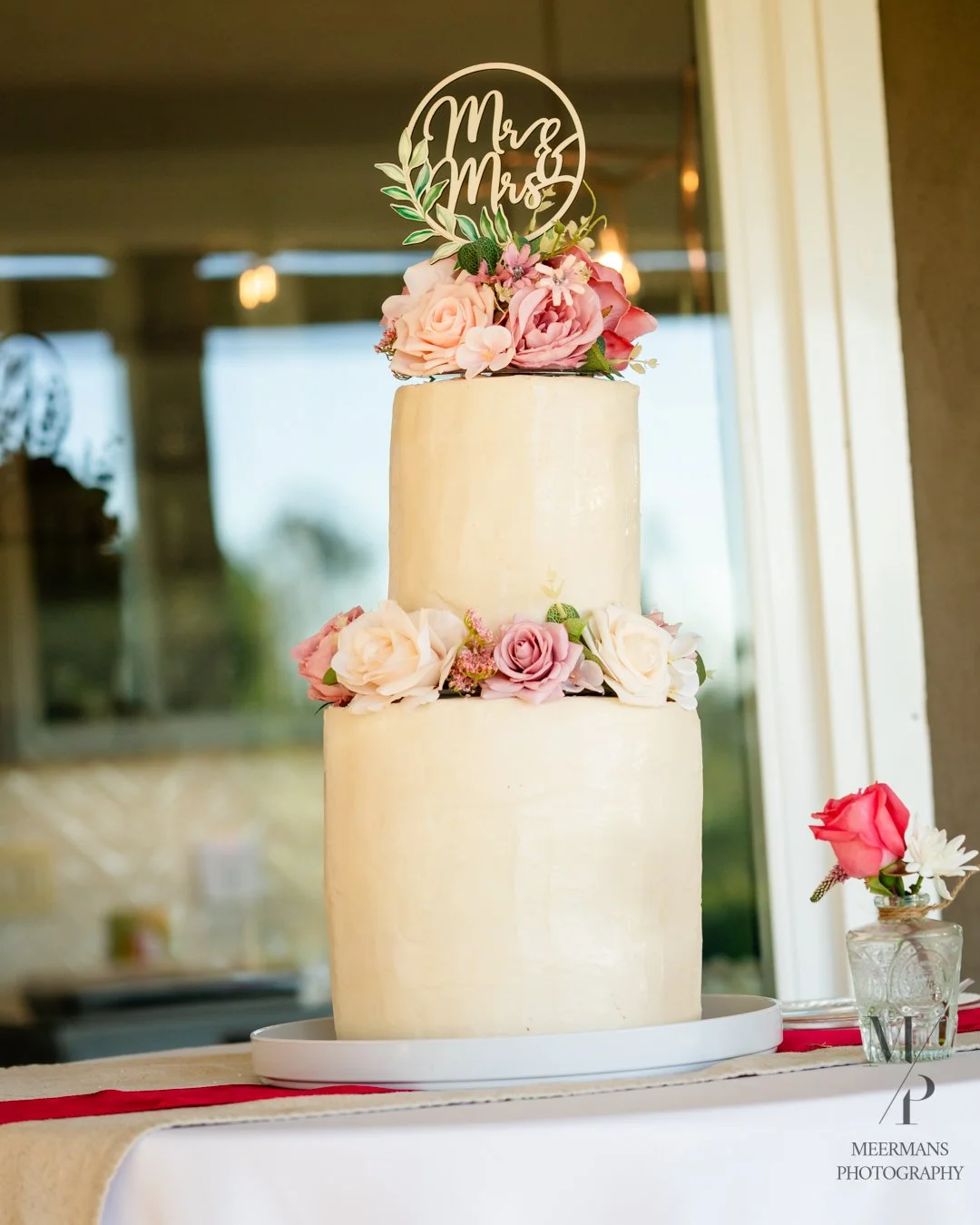 A three-tier wedding cake decorated with pink and white roses, topped with a cake topper that says 'Mr & Mrs', on a white cake stand with a small flower arrangement beside it.