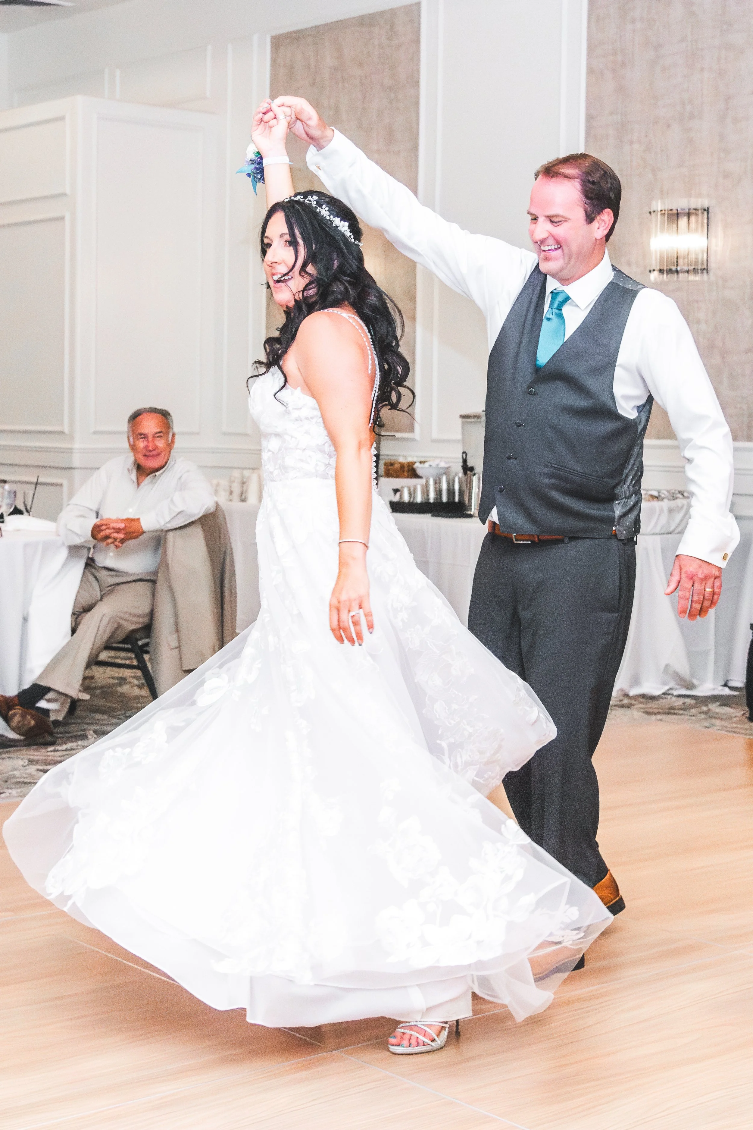 A bride and groom dancing at a wedding reception, with the bride twirling in her white gown and the groom smiling while holding her hand. A seated guest is watching and smiling in the background.