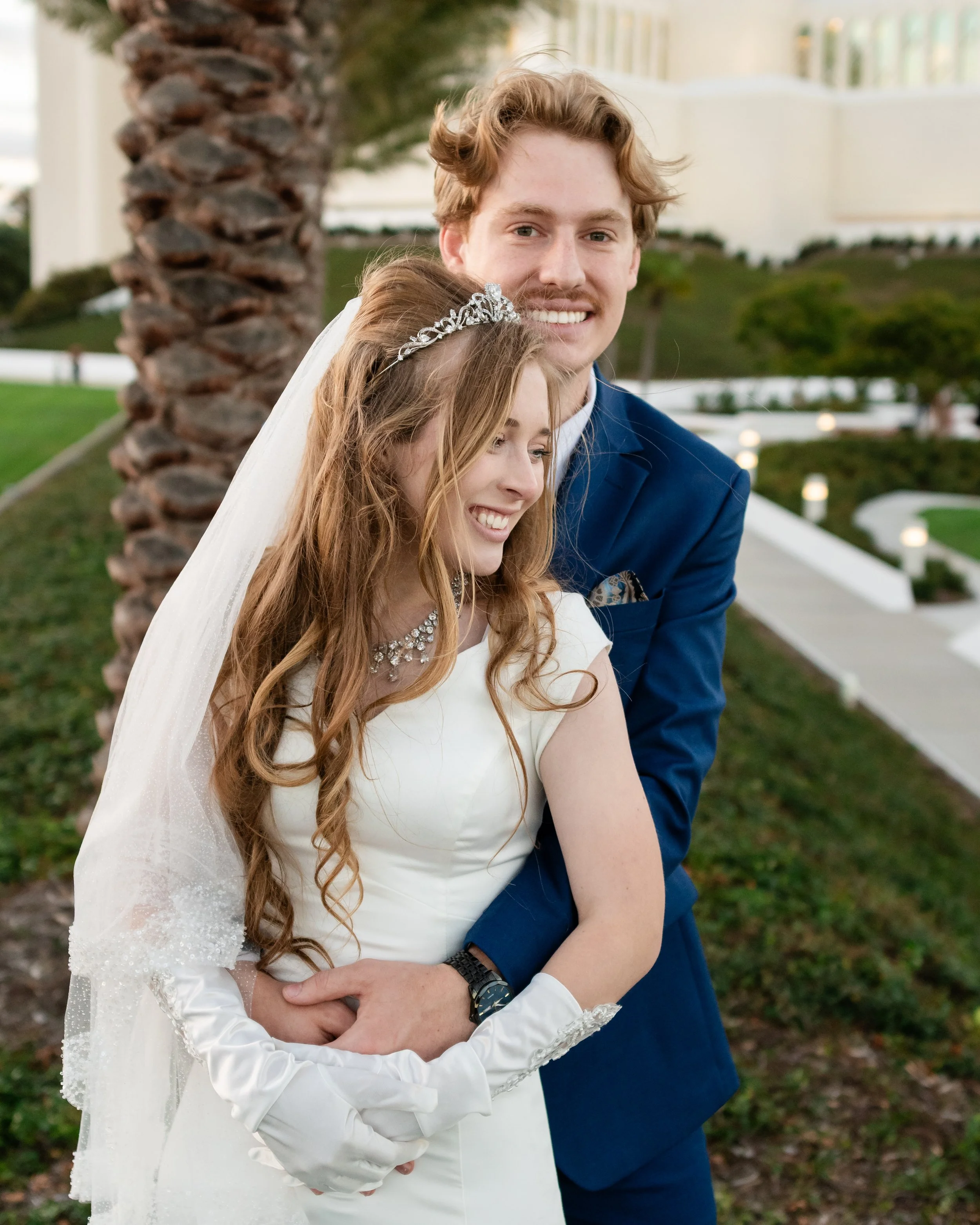 A couple dressed in wedding attire, standing outdoors near a palm tree, smiling, with greenery in the background.