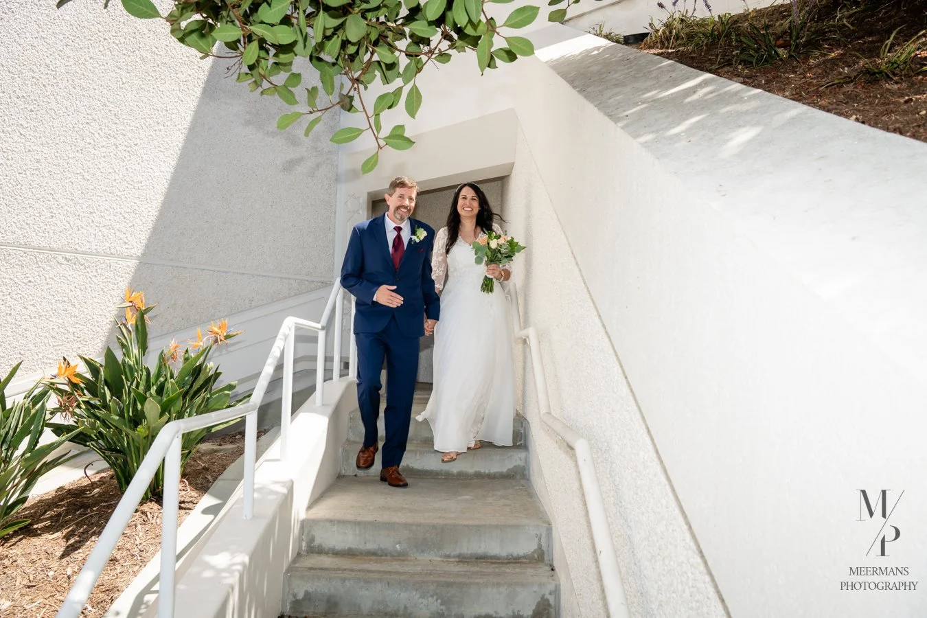 A newlywed couple smiling and holding hands on outdoor stairs, the bride holding a bouquet of flowers, with a white textured wall and plants nearby.