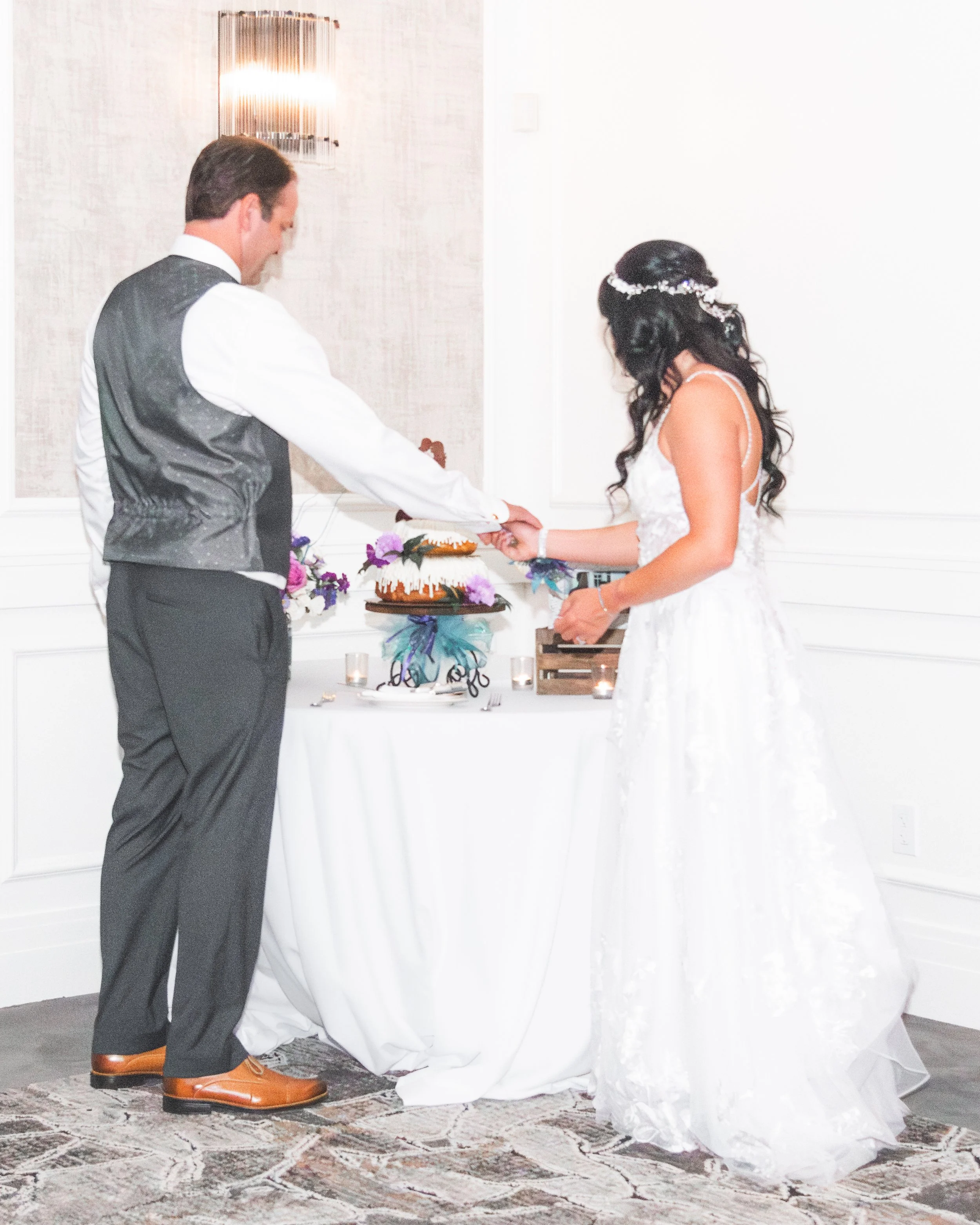 A bride and groom exchange wedding vows during their ceremony. The groom is dressed in a gray vest, white shirt, and brown shoes, kneeling on a rug. The bride is in a white wedding gown with dark hair and a floral headpiece. They hold hands over a ta