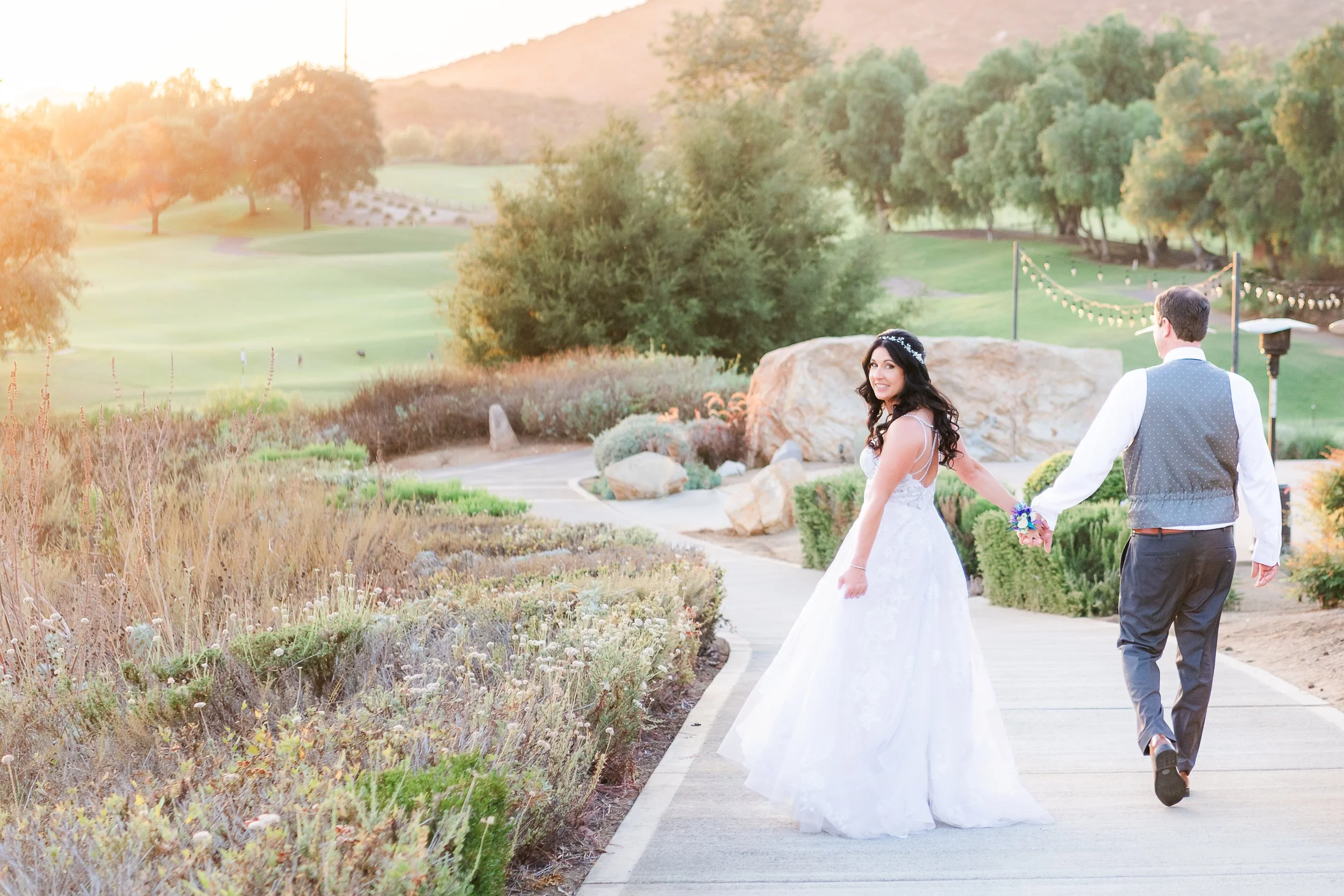 Couple dressed in wedding attire walking along a garden path during sunset. The bride is wearing a white wedding gown, and the groom is in a vest and jeans. The landscape features green fields, trees, and distant hills.