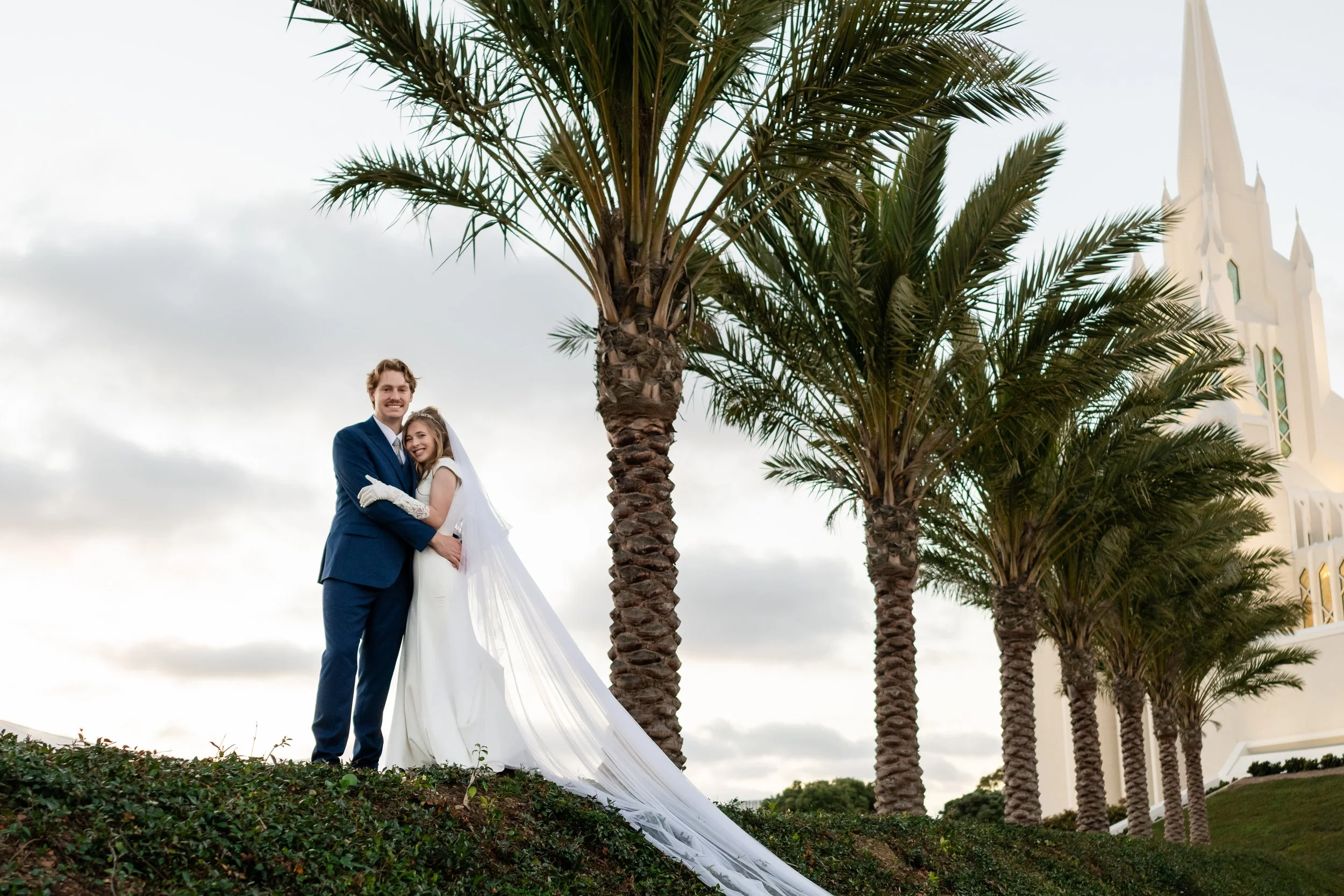 A couple in wedding attire standing outdoors among palm trees, smiling at the camera, with a church building in the background.