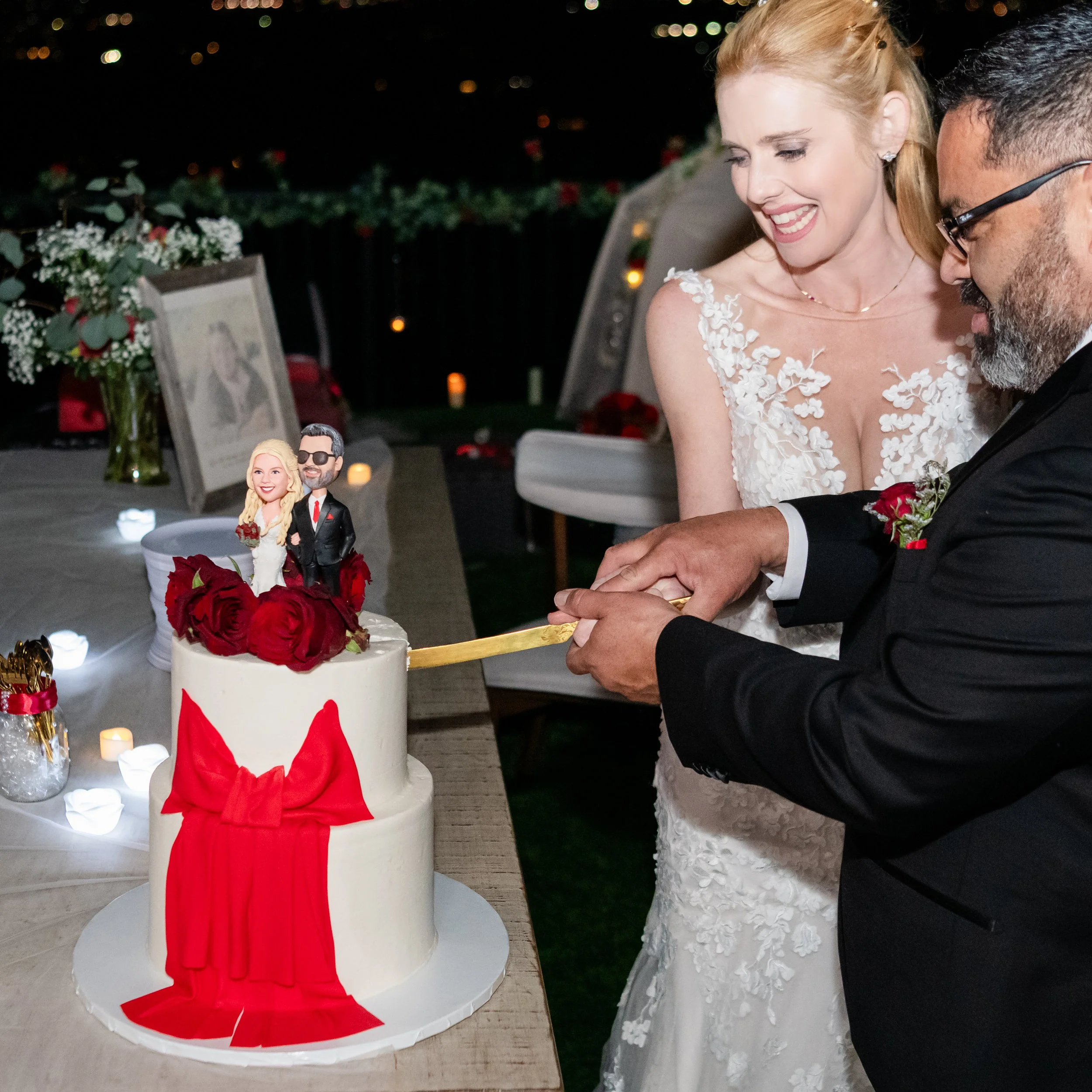 A woman in a wedding dress and a man in a suit are cutting a wedding cake with a knife. The cake is decorated with red roses and a bride and groom figurine on top, with candles and floral arrangements on the table.
