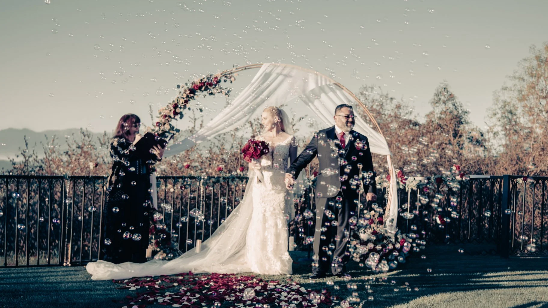 A bride in a white wedding gown and a groom in a dark suit hold hands during their outdoor wedding ceremony, surrounded by floating bubbles and rose petals, with an officiant reading from a book nearby.