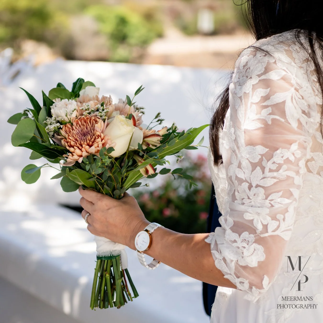 A woman in a lace wedding dress holding a bouquet of flowers, including white roses and peach-colored blooms, outdoors with blurred greenery in the background.