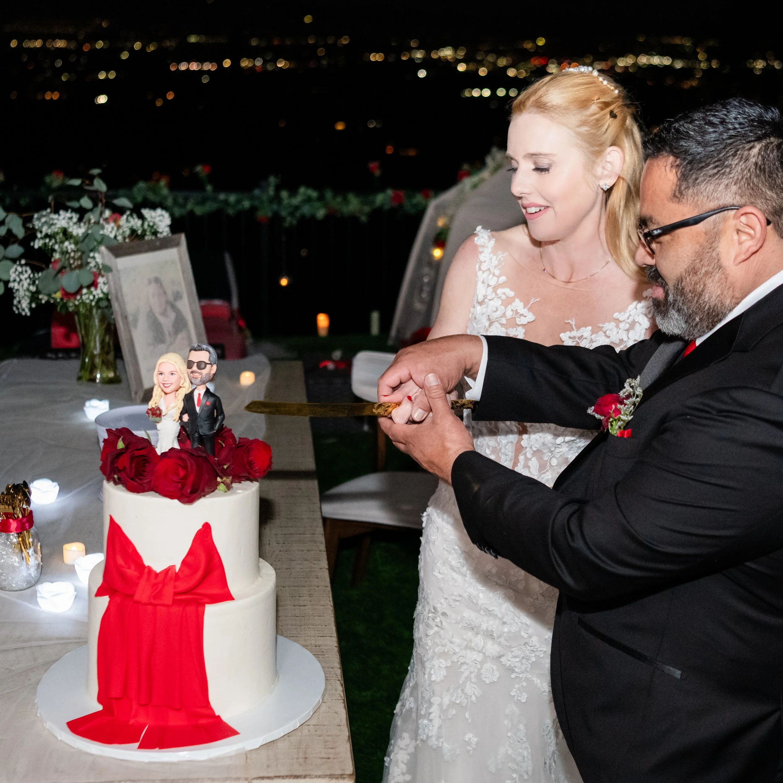 A bride and groom celebrating their wedding at night, with a cake decorated with red roses and a cake topper of a bride and groom figurine on top, and a city skyline in the background.