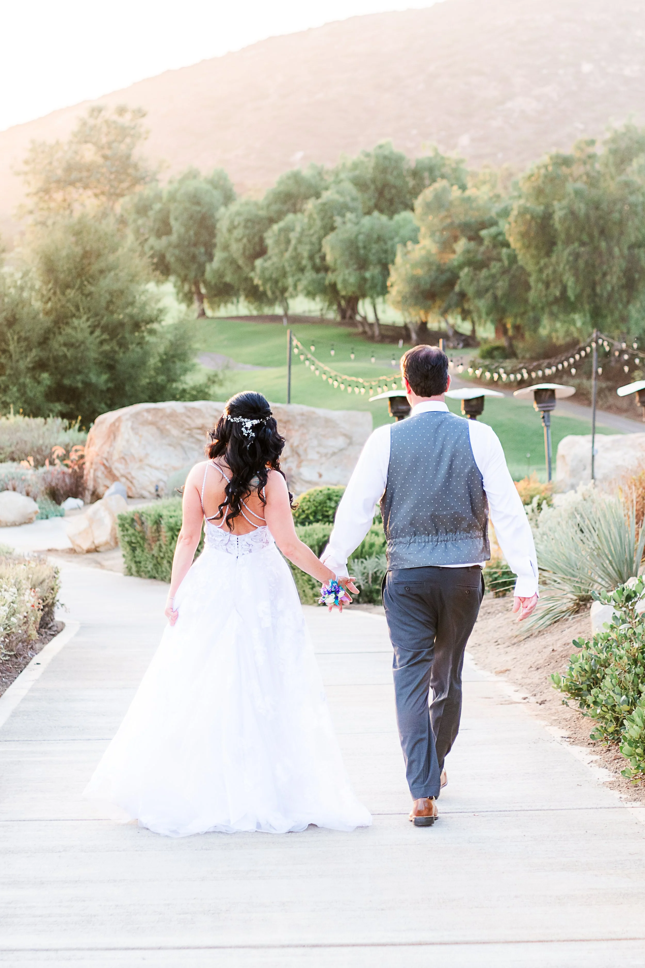 A newlywed couple walking hand in hand outdoors at sunset, with a scenic landscape of trees, hills, and string lights in the background.