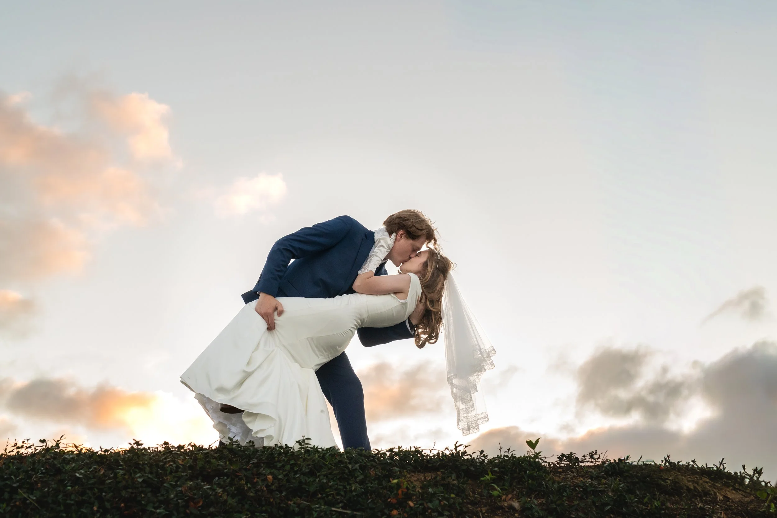 A bride and groom kissing during a wedding photo shoot outdoors with a sunset sky in the background.