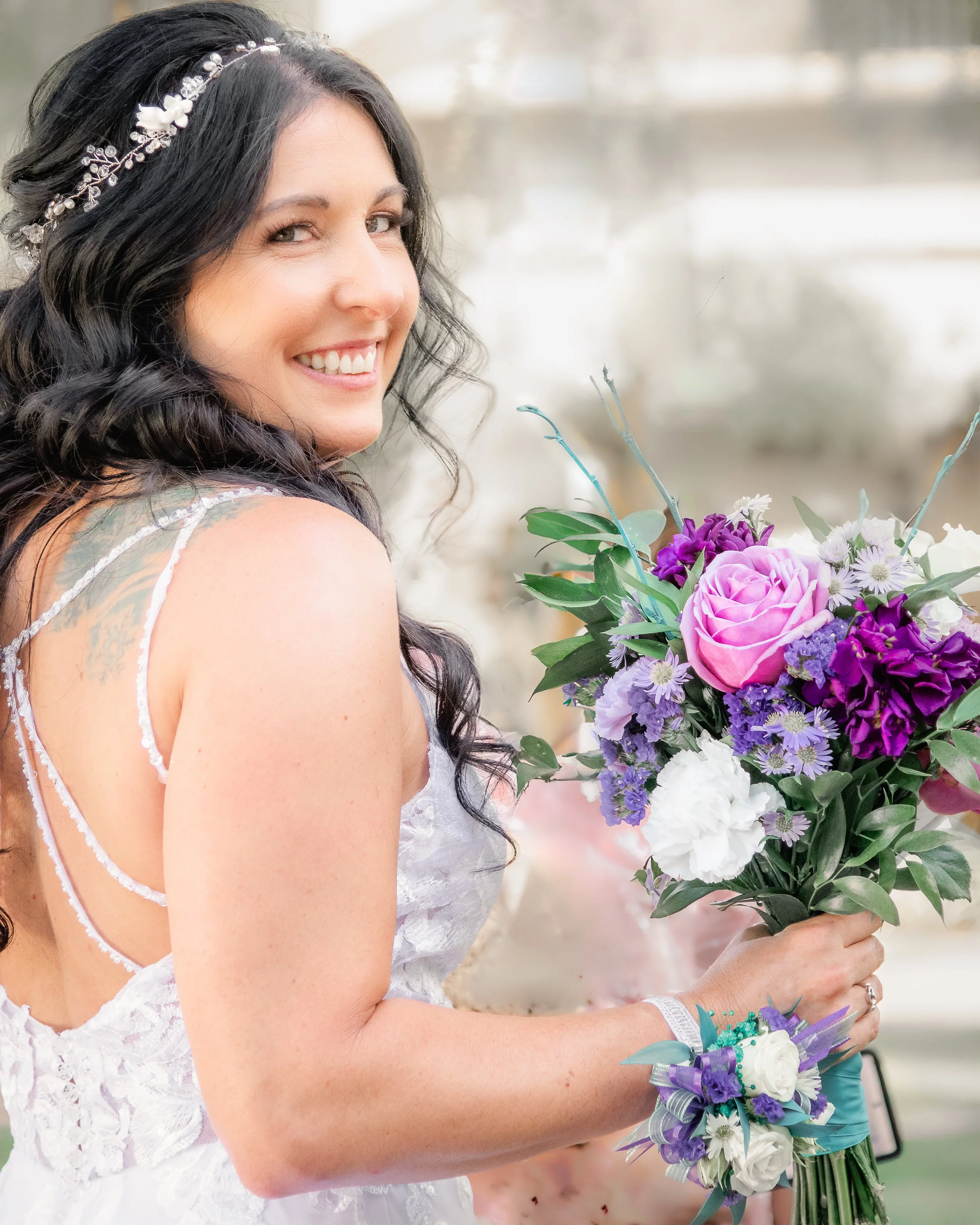 A bride with long dark hair holding a large bouquet of purple and white flowers, wearing a white lace wedding dress and a floral headband, smiling outdoors.